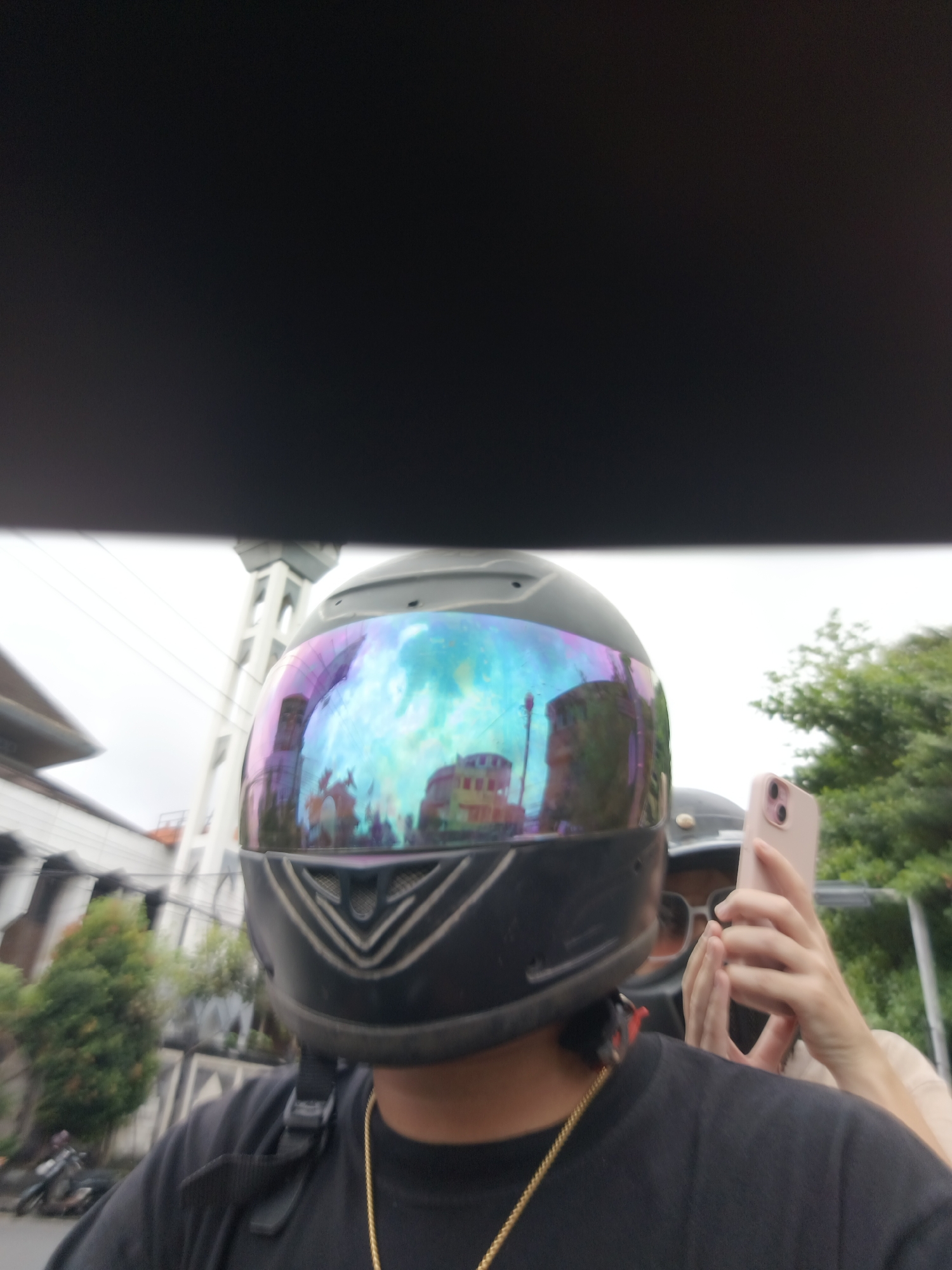 The image shows a person wearing a black motorcycle helmet with a colorful, iridescent visor. The visor reflects buildings and what appears to be a clear sky. The person is wearing a black t-shirt and a gold chain. In the background, there are trees and buildings, suggesting an urban or suburban environment. A hand holding a pink smartphone is visible on the right side of the image, partially obscuring another person's face. The general appearance of the surroundings, including the style of buildings and vegetation, is consistent with the city of Denpasar, Indonesia.