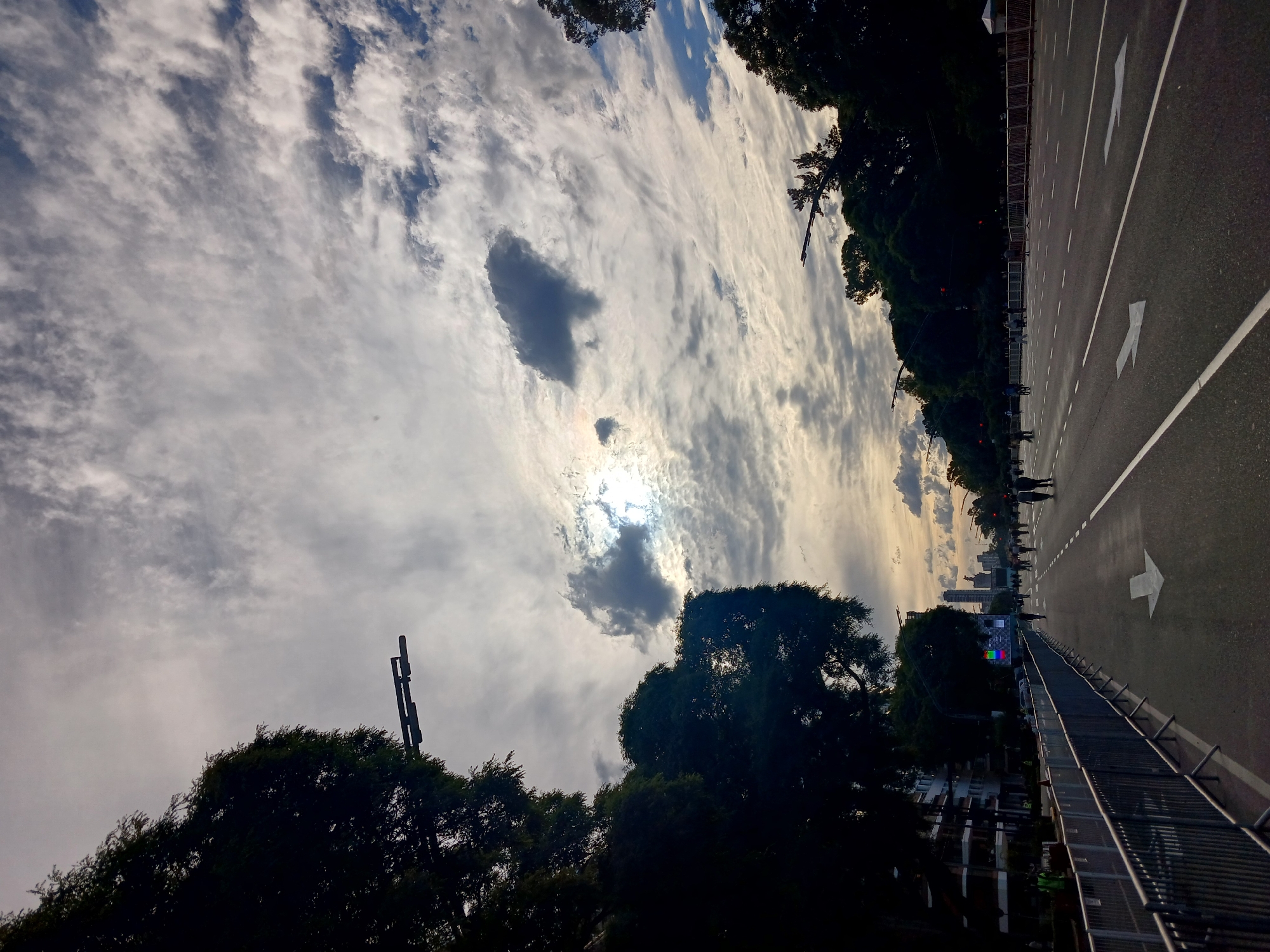 The image shows a street scene in Buenos Aires, Argentina, taken from a low angle. The sky is predominantly cloudy with the sun partially obscured. A paved road with white directional arrows and lane markings extends into the distance, flanked by metal barriers. A line of people can be seen walking along the side of the road. Trees and foliage line the side of the road on both sides. Buildings of the Buenos Aires skyline are faintly visible in the distance.