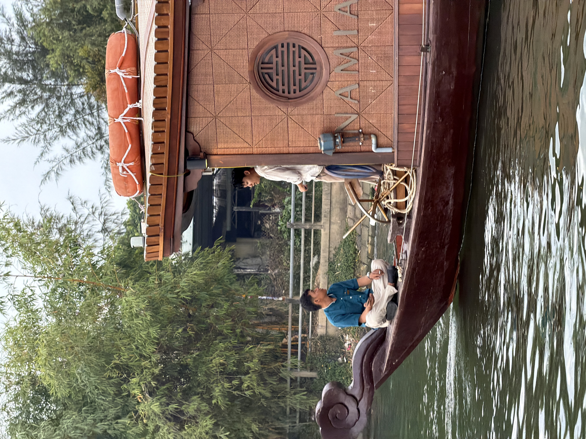 The image captures a traditional-style wooden boat on a calm body of water, likely a river or canal, set against a backdrop of lush green foliage and distant structures in what appears to be Đà Nẵng, Vietnam.
Two men are on board the boat. Towards the bow, one man, dressed in a teal or blue long-sleeved shirt and light-colored trousers, is seated with his legs crossed, looking towards the riverbank. He holds a small white object in his hands. Further back on the boat, another man, wearing a light-colored long-sleeved shirt and dark trousers, stands near a wooden steering mechanism, possibly operating the vessel or observing the surroundings.
The boat itself is dark brown with a richly textured, light brown cabin structure that features a distinctive circular, ornate wooden window with a traditional geometric pattern. The name "NAMIA" is vertically inscribed on the cabin's side. Two orange cylindrical life preservers are securely fastened to the exterior of the cabin. The bow of the boat is adorned with intricate dark wooden scrollwork.
The water is dark and reflective, showing gentle ripples. The riverbanks are densely covered with green, bamboo-like trees. In the background, a metal railing and some indistinct structures can be seen. The sky appears overcast, indicating daytime with soft, diffused lighting. The scene suggests a peaceful journey or tour on the water.