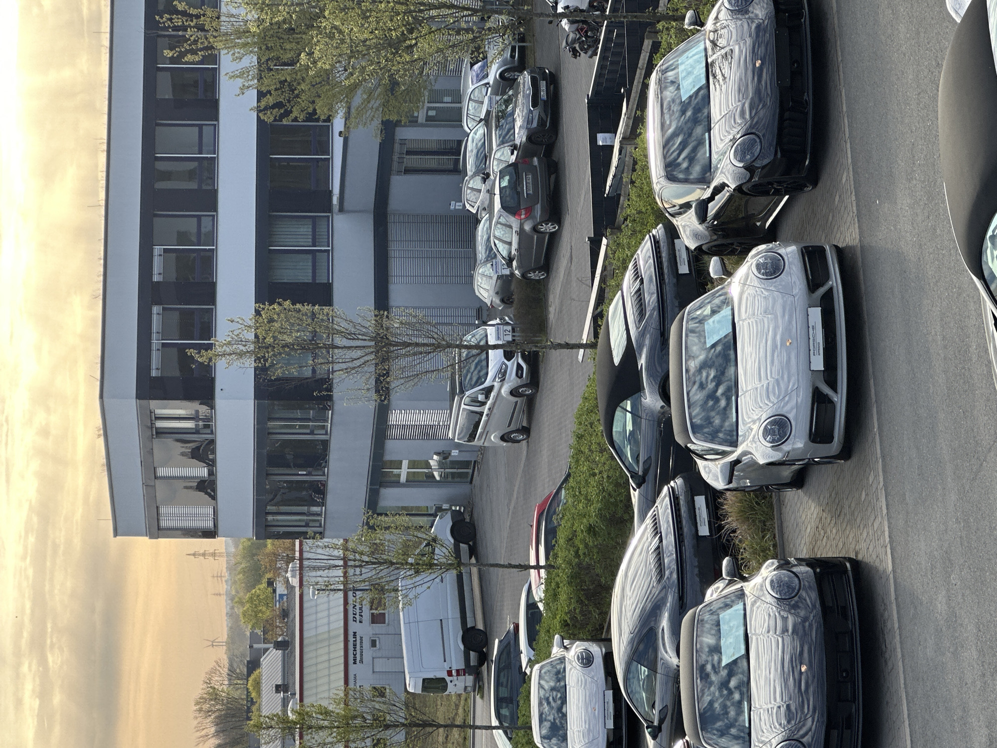 The image shows an outdoor scene in Holzwickede, Germany. A modern, multi-story building with a light gray facade and dark window frames is prominently featured. In the foreground, there is a row of parked cars, appearing to be new Porsches, mostly in shades of gray and black. A small tree is planted between the vehicles and the building. In the background, across the street, another building is visible. This building has signage indicating the presence of Michelin, Dunlop, and other tire-related businesses. A white van is parked near the signage. Utility poles and what appears to be a wind turbine are distantly visible against a yellowish sky, possibly indicating early morning or late evening light. The overall impression is of a commercial area with a focus on the automotive industry.