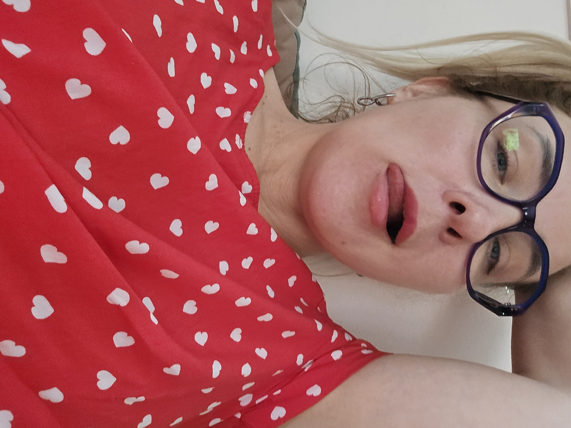 A fair-skinned woman with blonde hair is lying down, facing the camera at an angle. She wears glasses with blue frames. Her red dress is patterned with small white hearts. The background is a plain, light surface. No information relates the image to the city of Sochi, Russia.