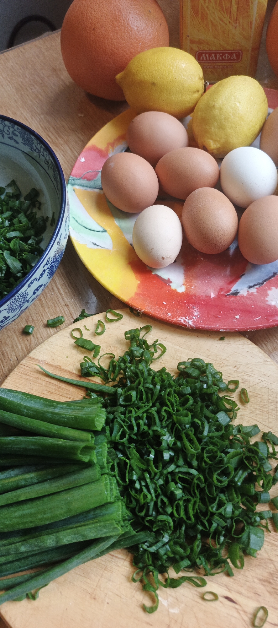 The image shows a close-up, overhead view of a wooden table with various food items. In the upper left is a grapefruit, and to its right are two yellow lemons. Below the grapefruit and lemons is a colorful plate with several eggs, some brown and one white. To the left of the plate, a blue and white patterned bowl contains chopped green onions. In the foreground, a wooden cutting board is covered with a pile of chopped green onions, and a bunch of whole green onions is to the left. The background shows a yellow box with Cyrillic lettering, and the wooden surface of the table.