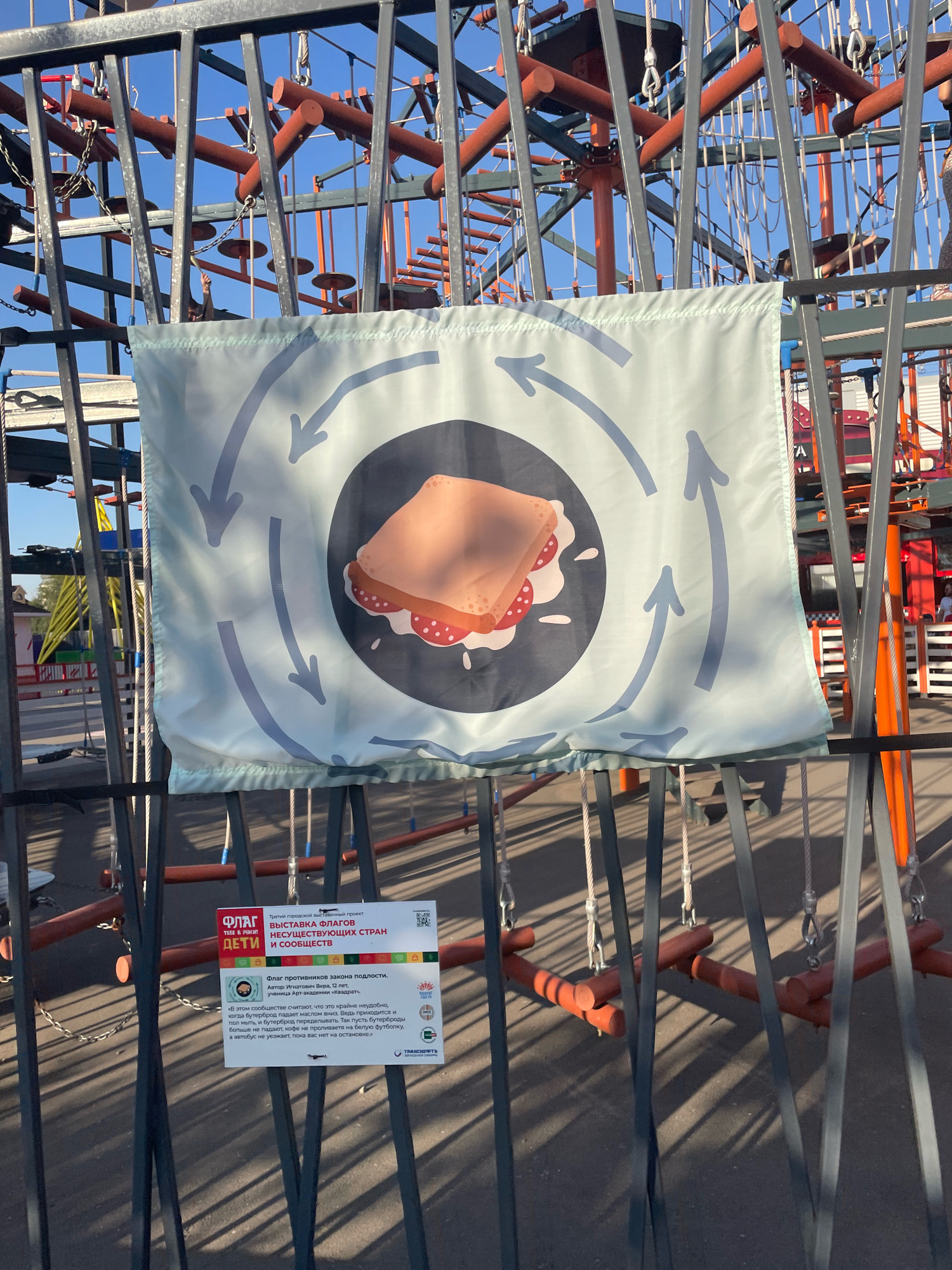 The image shows a close-up view of a section of an outdoor ropes course or adventure park. In the foreground, a light blue flag with a circular graphic depicting a sandwich surrounded by swirling arrows is attached to a metal fence. The flag appears to be part of an exhibition or event. Below the flag, a white informational sign is also attached to the fence. The sign features text in Russian, including "ФЛАГ" (FLAG), "ВЫСТАВКА ФЛАГОВ" (FLAG EXHIBITION), and "ДЕТИ" (CHILDREN), along with a QR code and some small colorful icons. The ropes course structure, made of metal beams and wooden elements, is visible in the background, along with cables and ropes. The ground beneath the structure appears to be asphalt. The lighting suggests it is daytime, with shadows cast by the structure on the ground. This event is identified as part of the "Тайны городской проект" (Secrets of the City Project) and specifically mentions the city of Omsk, Russia, through its logo which includes a stylized image of the Omsk Kremlin and a heraldic symbol.