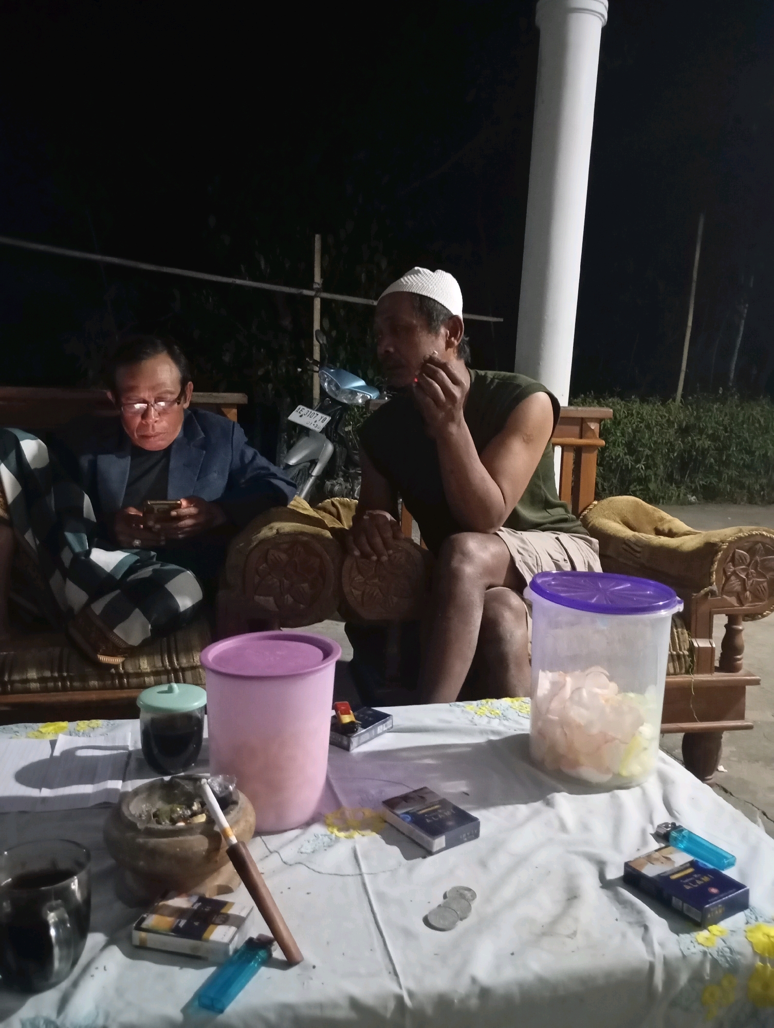 A nighttime outdoor scene captures two men sitting on a sofa. The man on the left wears glasses and a suit jacket over a black shirt, engrossed in his mobile phone. He is positioned on a striped cushion with a checkered cloth draped over his lap. The man on the right, wearing a white skullcap and a dark green t-shirt with shorts, is smoking a cigarette. He sits with his legs crossed, his right arm resting on his knee, holding the cigarette. In the background, a white pillar stands vertically, and a motorcycle is visible.
In the foreground, a white tablecloth is spread over a table. On the table, various items are arranged: a stone ashtray with a burning cigarette, a pack of cigarettes, a glass of dark liquid, a small green-lidded container, a large pink lidded plastic container filled with white and pink translucent items, a blue lighter, and several loose coins. The lighting suggests it is nighttime, with the scene illuminated by ambient light sources, possibly from nearby structures or the flash of the camera. The overall environment appears to be a residential outdoor area, possibly a porch or patio. The presence of a motorcycle and the casual seating arrangement suggest a relaxed, social gathering.