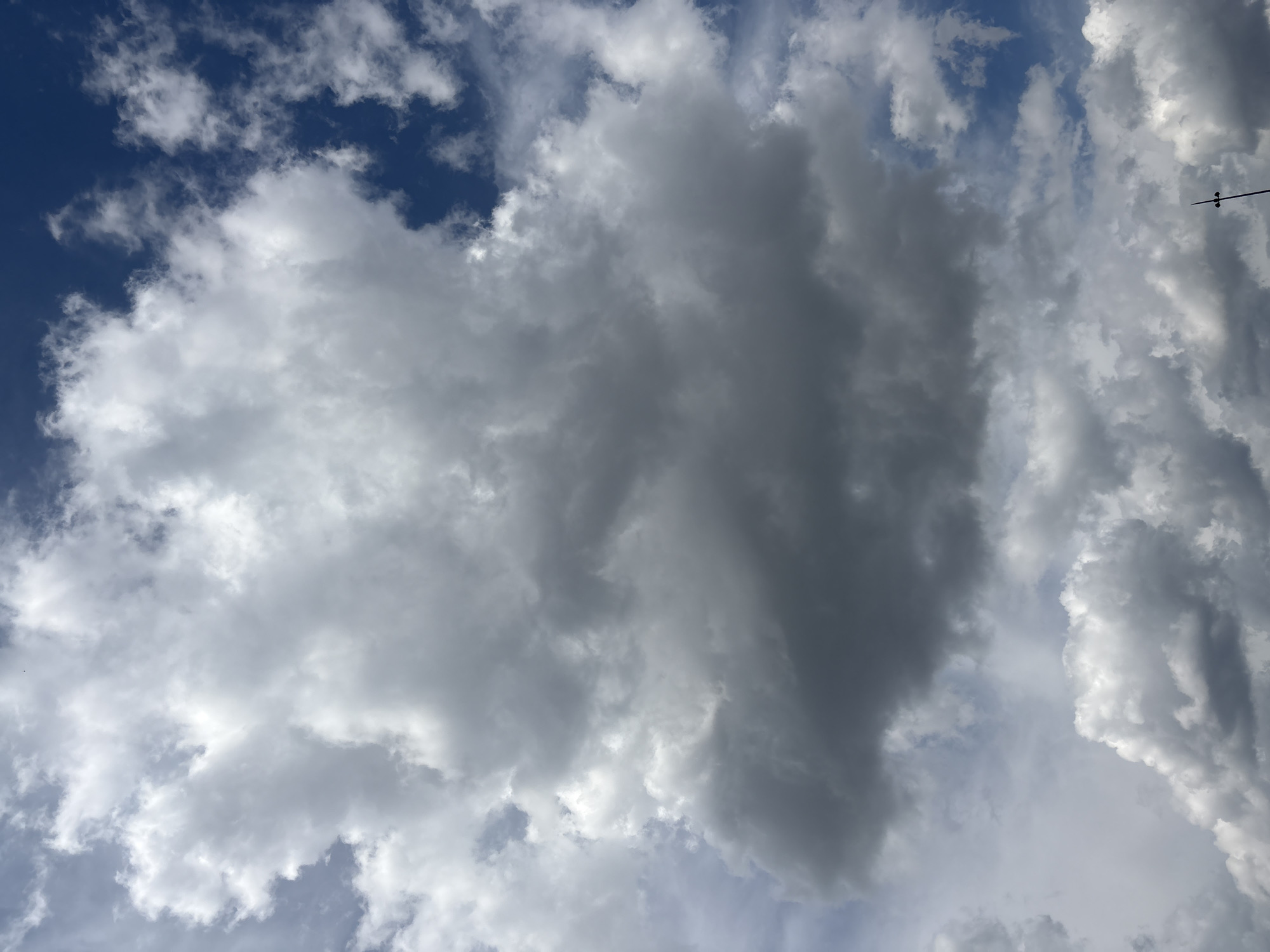 The image is a close-up, vertical shot of a turbulent sky filled with dramatic clouds against a bright blue sky. The clouds vary in shade from bright white where the sun hits them to dark gray in their shadowed undersides, suggesting impending weather changes. In the bottom right corner, a small portion of a tall, dark pole with two small spherical lights or antennas is visible, possibly a lamppost or a street signal. There are no people or discernible city structures visible, indicating the photo was taken from a vantage point where the sky is the primary focus, such as a rooftop or an open area. The scene is devoid of any specific Moscow landmarks or identifying features, making it impossible to determine its precise location within the city.