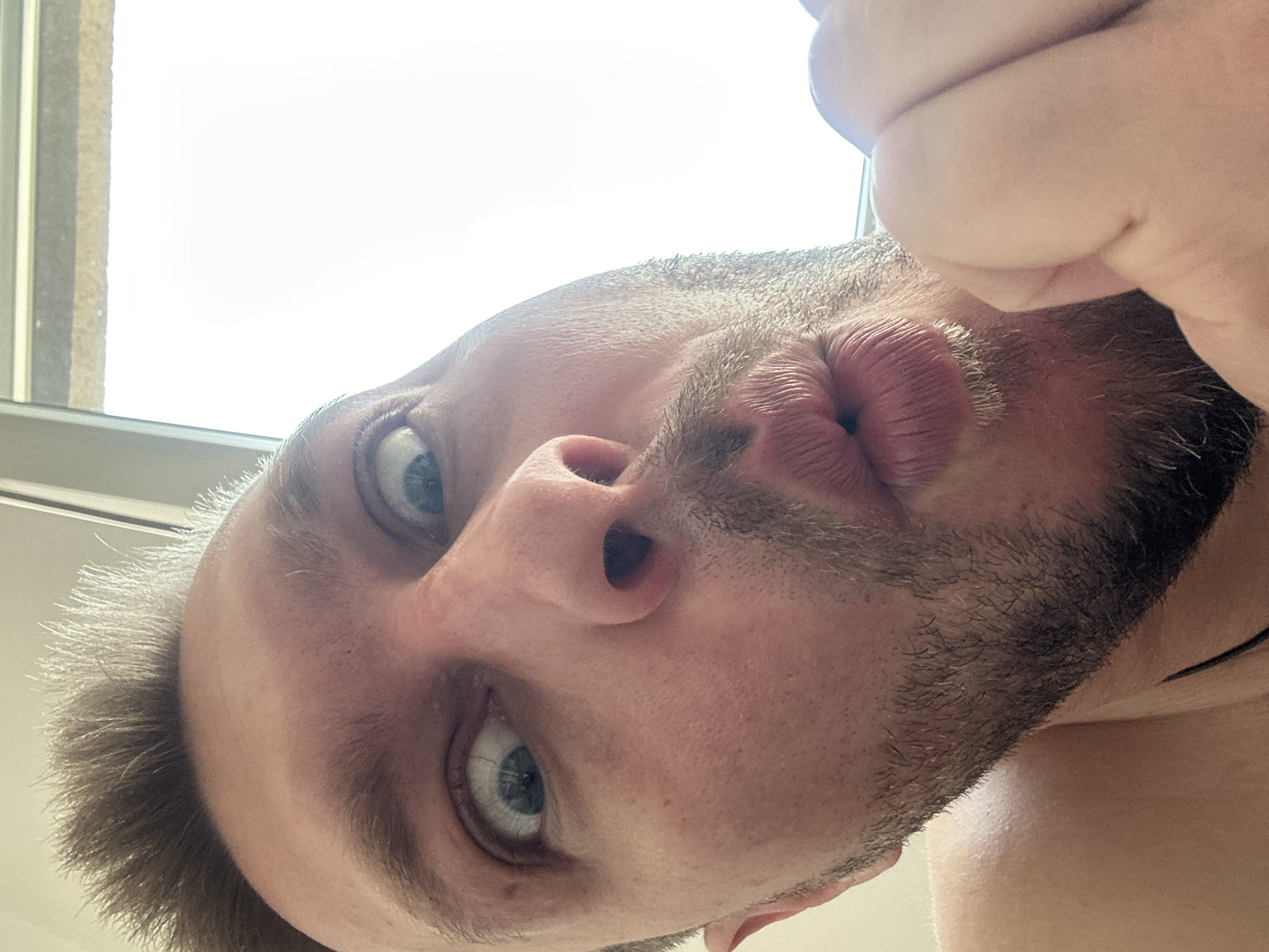 The image is a close-up, high-angle, selfie-style shot of a person's face. The person has light skin, brown hair styled upwards, and light blue eyes. They are making a puckering or kissing face with their lips pursed. There is stubble visible on their chin and upper lip. Their right hand is held up near their mouth. The background is bright, suggesting it is daylight, possibly near a window. The scene is not identifiable as being in Zvezdara Urban Municipality, Serbia.