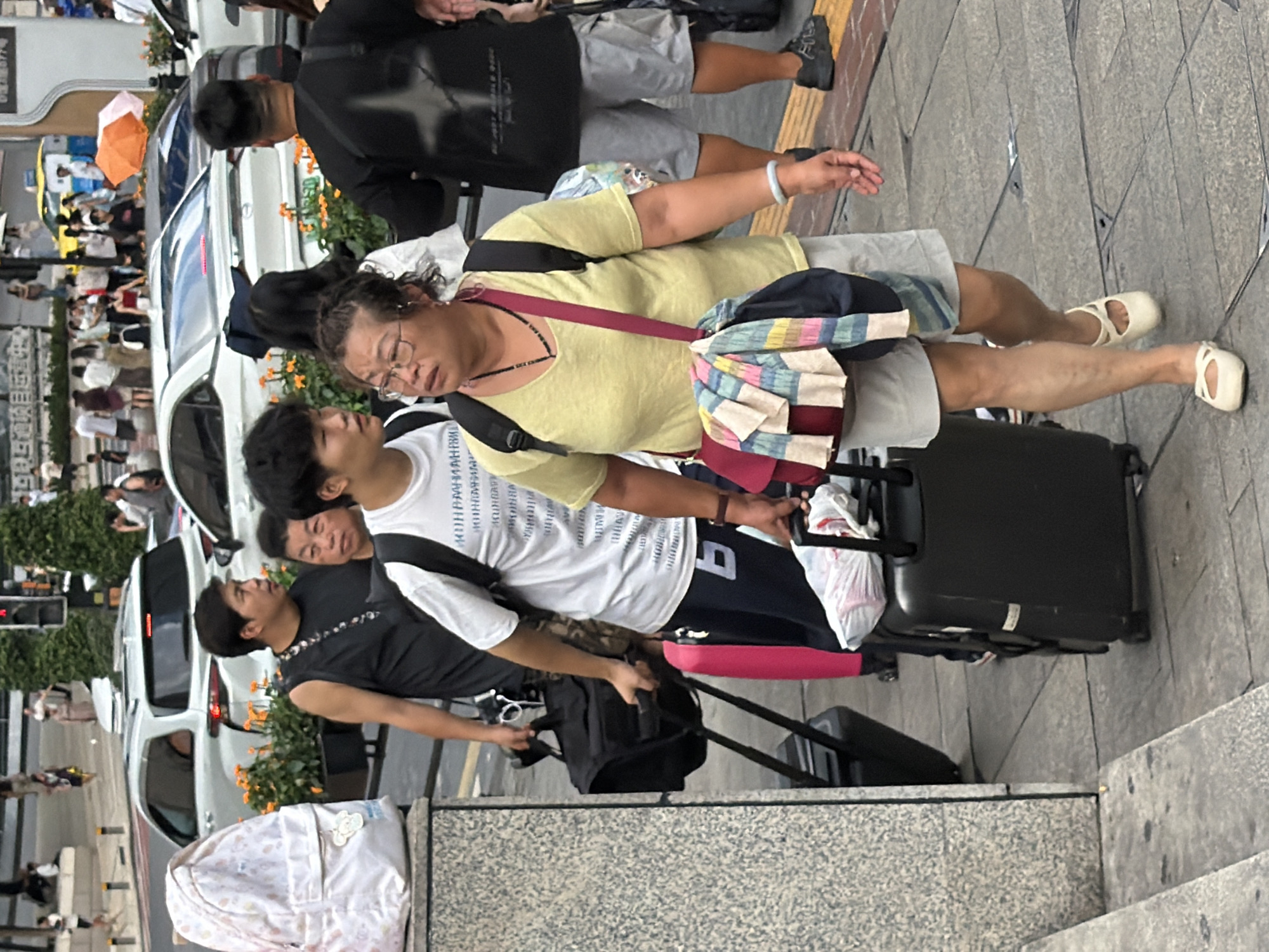 In Yuzhong District, China, a group of people are seen navigating a sidewalk with luggage. In the foreground, a woman wearing a yellow t-shirt, glasses, and light-colored shorts walks with a rolling suitcase. A colorful striped cloth is draped over her shoulder bag. To her left, a young man in a white t-shirt and dark pants walks alongside her, also with a backpack. Behind them, a man in a black sleeveless shirt is pulling a black rolling suitcase, and another man in a black t-shirt and grey shorts walks past. In the background, white cars are parked, and several people are visible on the street and sidewalk, suggesting a busy urban environment. The scene is captured on a daytime, possibly overcast day, given the lighting.