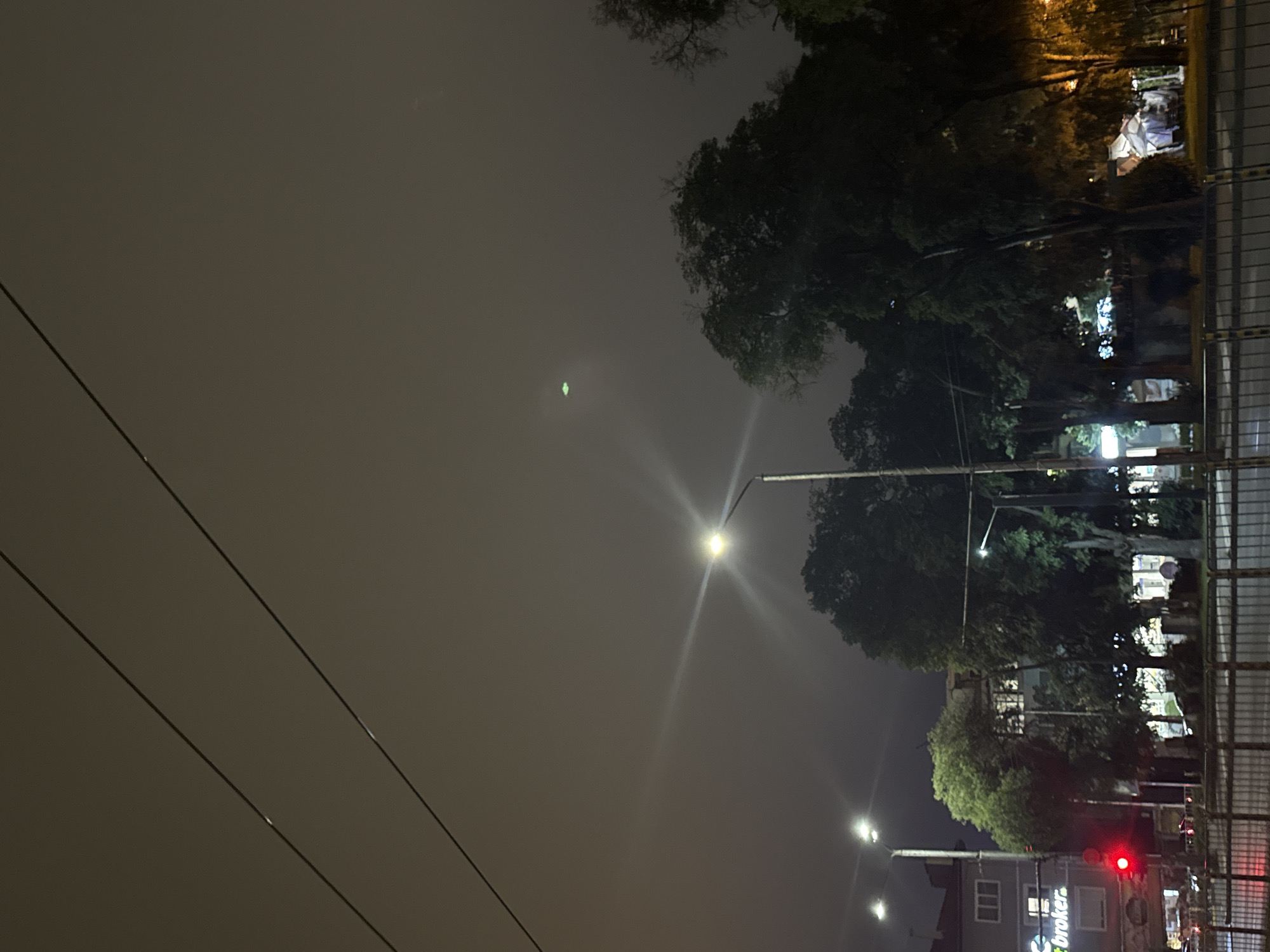 The image captures a nighttime urban scene, likely an intersection adjacent to a public green space, under an overcast or foggy sky. Two parallel, dark overhead wires traverse the upper third of the frame. High in the sky, a distinct, oval-shaped green light source is visible through the atmospheric haze.
In the foreground, a dark metal fence or railing runs horizontally across the lower portion, separating the viewer's perspective from a wet or reflective paved surface, possibly a street.
On the left side of the image, a light-colored multi-story building with dark window frames is partially visible. The ground floor of this building features a green illuminated sign with partial text reading "t broker." A red traffic light is active and prominently displayed. Multiple streetlights illuminate the scene, casting bright white or yellow light and producing prominent lens flare effects, particularly from a central streetlamp.
The center and right side of the image are dominated by a dense cluster of trees with dark green foliage, suggesting a park or a heavily treed median. Through the trees on the far right, a large, angular, light-colored structure, possibly a temporary installation or tent, is partially visible and appears illuminated. No individuals are discernible within the frame.
The location is in the city of Unknown, Unknown.