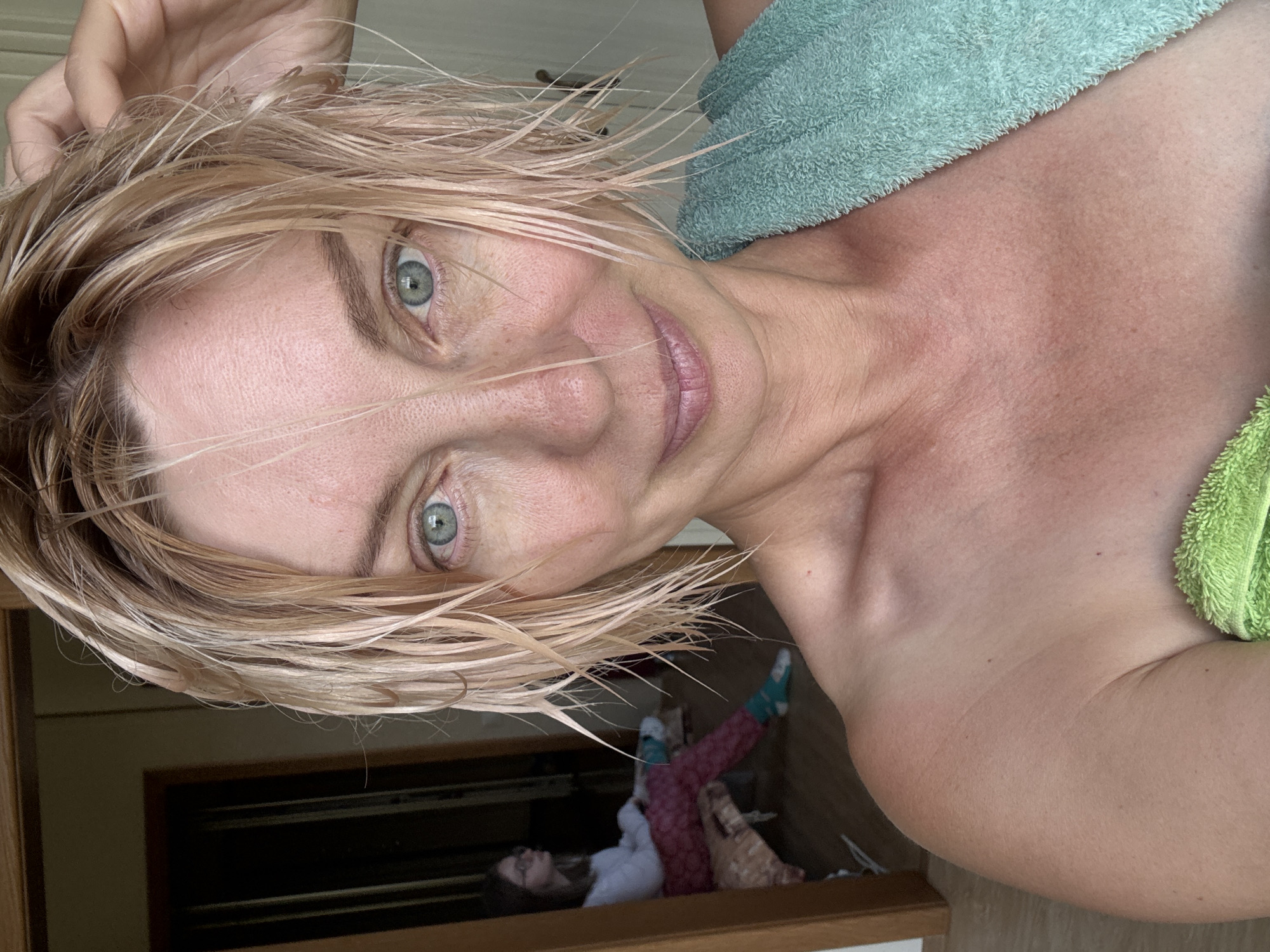 The media file is a portrait-oriented selfie taken by a woman with wet, blonde hair and blue eyes. She is wearing a green towel draped around her shoulders. Her skin appears flushed, and her lips are slightly parted, giving her a relaxed or perhaps slightly surprised expression. The background is blurred but suggests an indoor setting, possibly a bathroom or bedroom, with wooden flooring visible.
In the lower portion of the image, a child is visible lying down, seemingly on the floor or a low surface. The child is wearing pajamas and has glasses. This suggests a domestic scene, and the presence of two people, a woman and a child, indicates a familial context.
The lighting appears natural, suggesting it could be daytime. There are no visible textual elements in the media file. The woman's hair is damp, indicating she may have recently showered or bathed. The overall mood of the image is intimate and casual. The location context of Sochi, Russia, doesn't provide specific visual cues within the image itself.