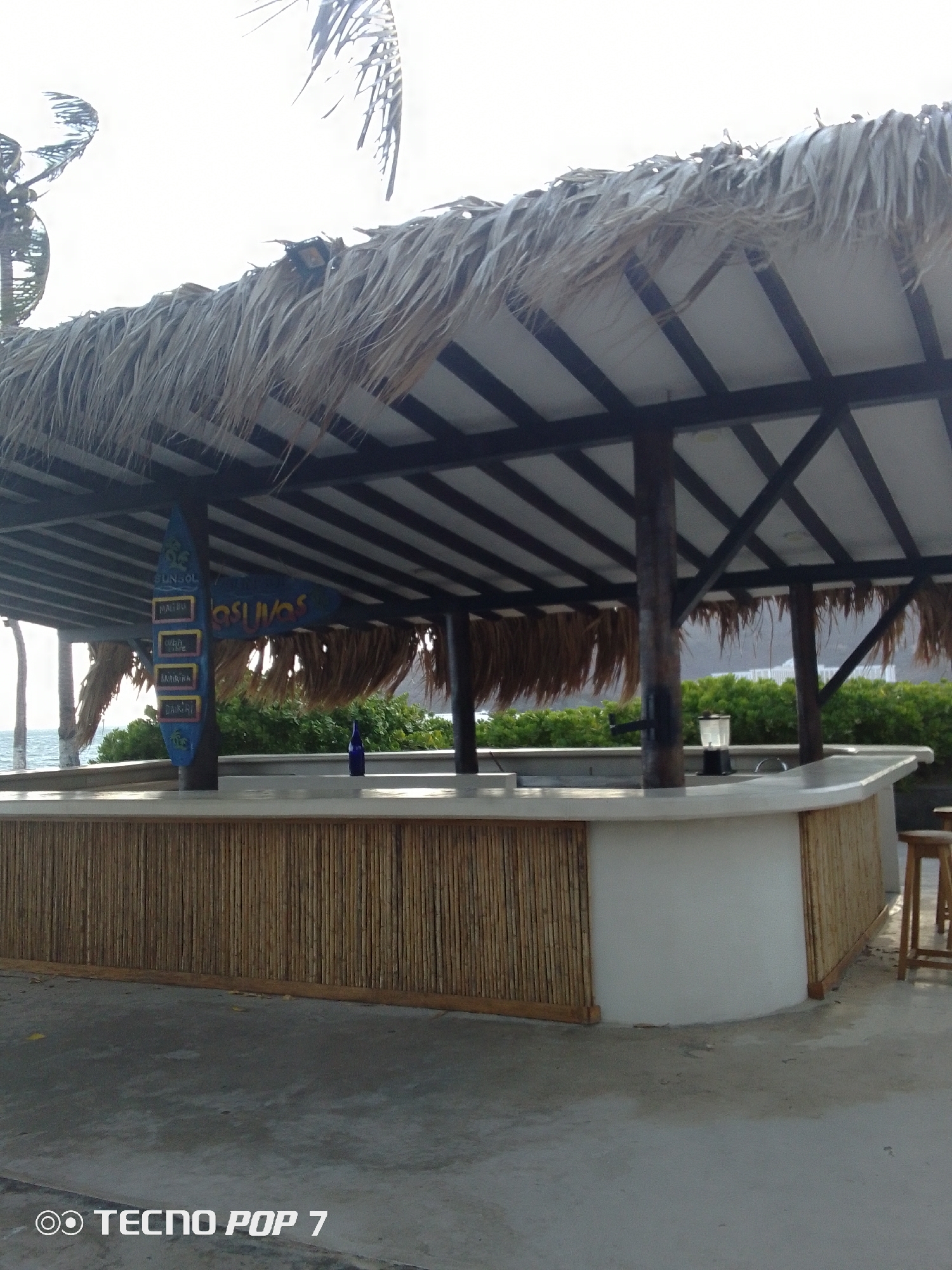 The image shows a partially enclosed outdoor bar in Pedro González, Venezuela. The bar is under a thatched roof supported by dark wood beams. A sign with the name "LAS UVAS" is visible along with a vertical sign shaped like a surfboard listing drink options. The bar's front is a combination of light-colored bamboo paneling and white stucco. A bottle sits on the bar top. A blender is seen behind the bar. A wooden stool sits at the bar's right side. The floor is concrete, with a phone model "TECNO POP 7" watermark visible in the lower left corner. The bar is set against a backdrop of green shrubbery and the ocean.