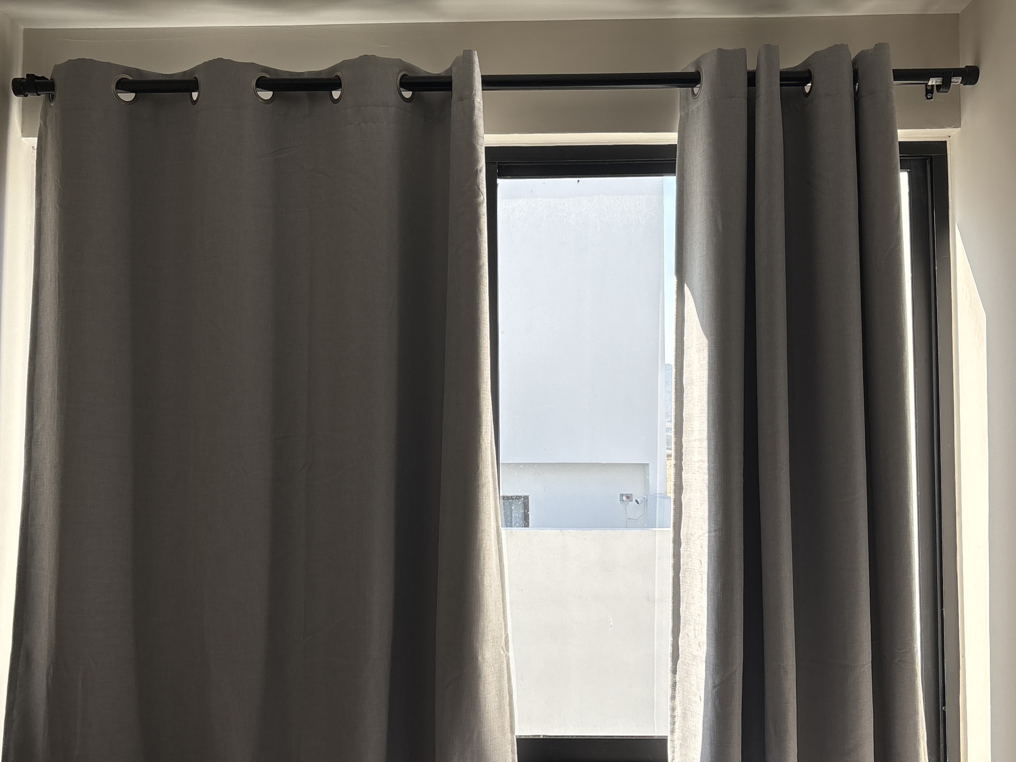 The image shows a window with gray curtains in what appears to be an interior space. The curtains are hung on a black rod with visible metal grommets. Through the window, a white wall is visible, suggesting an outdoor or another building. The lighting indicates that the photograph was taken during the day. The image does not provide enough information to determine the specific location in Santa Clara Ocoyucan, Mexico, or any details about people, subjects, objects, actions, or interactions beyond what is explicitly shown.