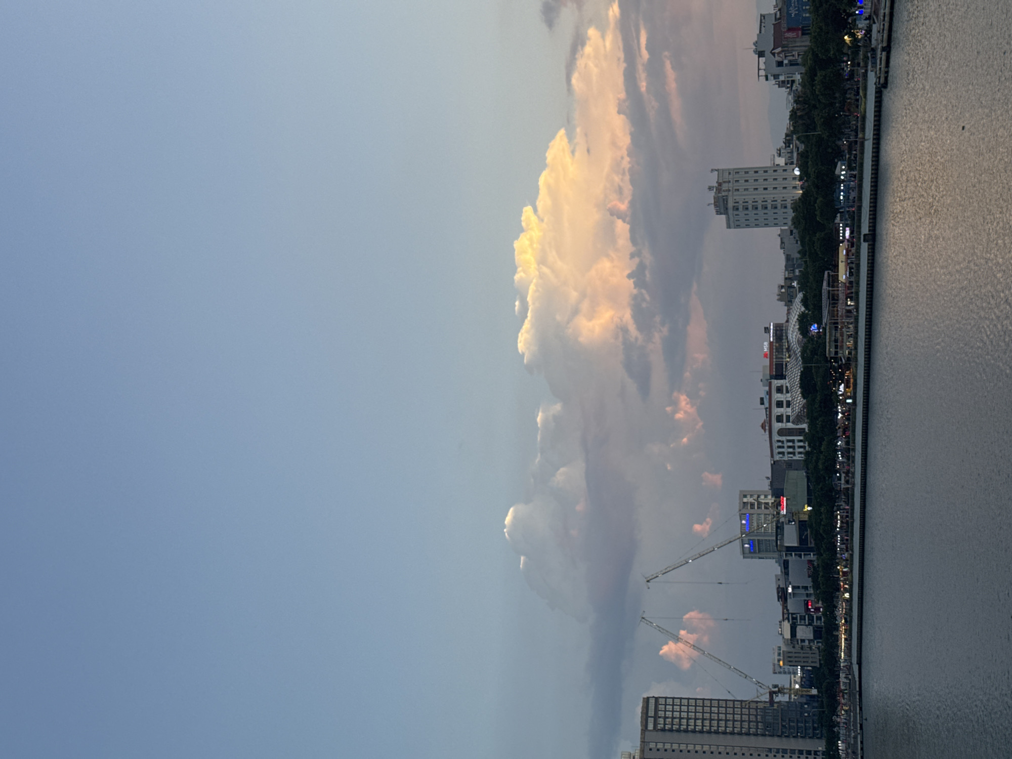 The image shows a cityscape in Đà Nẵng, Vietnam, at dusk. A large cumulonimbus cloud, illuminated by the setting sun, dominates the sky. The cloud is positioned above the city's skyline, which includes several high-rise buildings, construction cranes, and a Ferris wheel. A body of water, likely the Han River, runs along the edge of the city, reflecting the ambient light. The city's waterfront is lined with trees and streetlights.