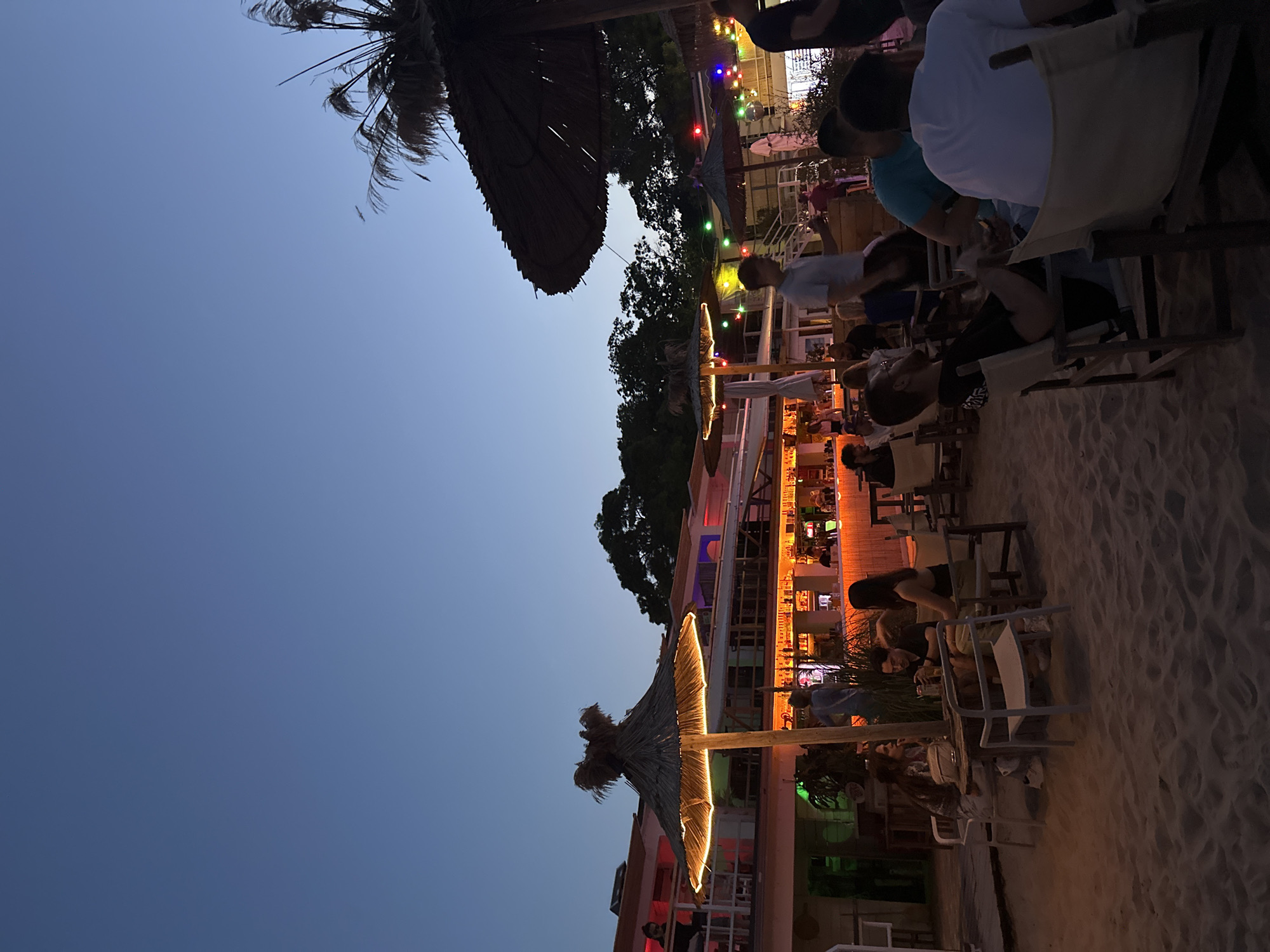 The image captures a scene at a beachside establishment in Varna, Bulgaria, during twilight. The setting is a sandy area with several groups of people seated at tables, suggesting a dining or bar environment. The architecture features multiple levels with open-air seating, and straw-thatched umbrellas provide shade over some tables. Decorative string lights are visible, contributing to the ambiance. A prominent bar area is illuminated with colorful lights, indicating it is operational. The sky is a deep blue, transitioning from daylight to night. The overall impression is of a lively evening gathering at a beach resort or restaurant.