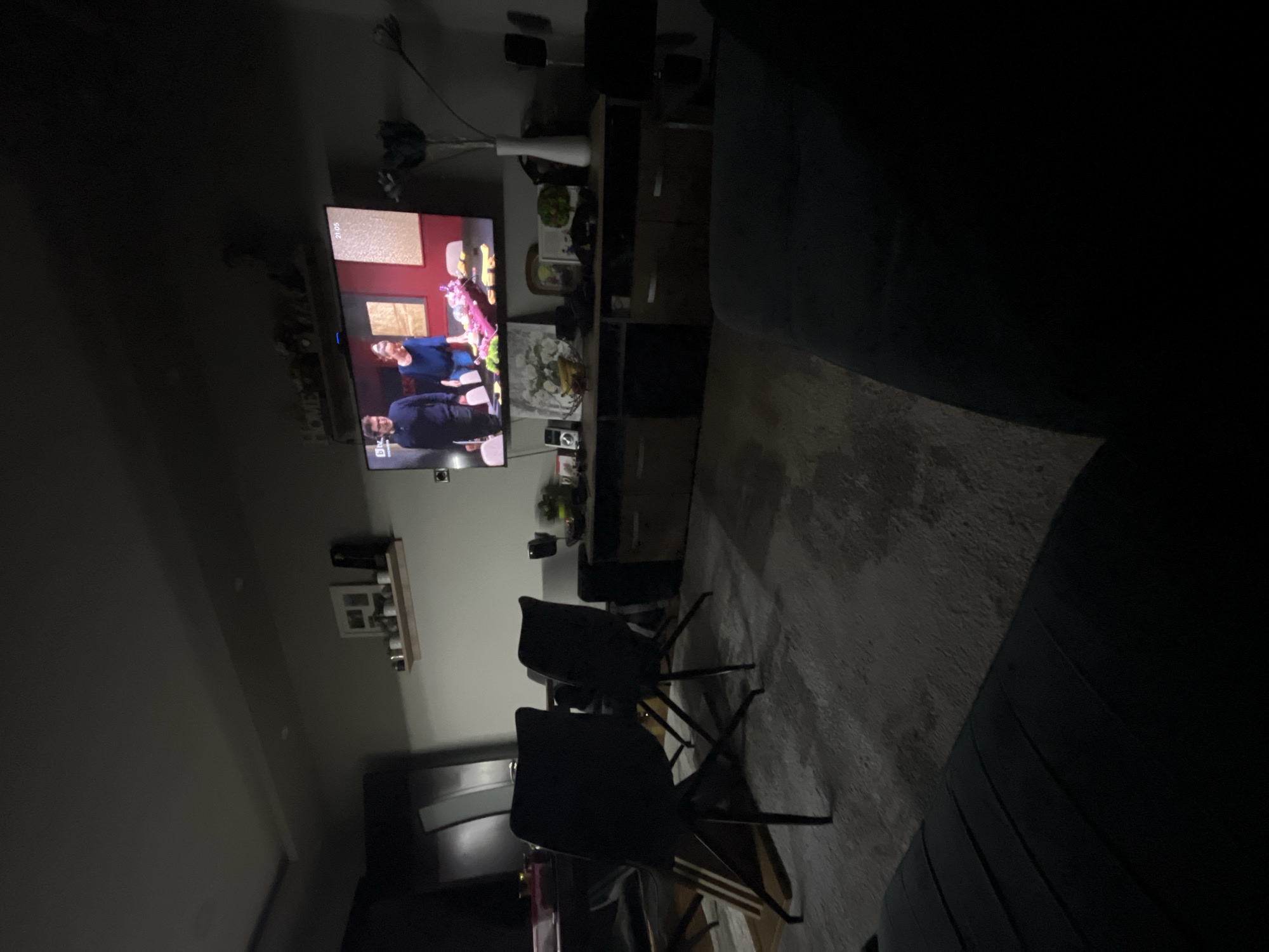 The image depicts a living room at night. A television displays a scene with two individuals, a man in a dark suit and a woman in blue. In front of the TV are two black chairs positioned on a rug. Next to the television is a shelving unit. A couch is visible on the right side of the frame.
There is no evidence of the location being Burgas, Bulgaria.