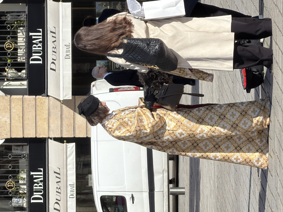 The image captures an outdoor urban scene in Place Vendôme, Paris, France, identifiable by the prominent "DUBAIL" luxury jewelry store signage on a classic stone building facade in the background. In the foreground, two female figures are positioned. On the left, a female with short hair wears a black hat with a bow, black gloves, and a long, intricately patterned gold and white brocade coat; she is looking downwards and holding a dark brown leather handbag. To her right, a second female figure is seen from the back, with long brown hair, wearing a light beige long coat and black wide-leg trousers; she carries a black quilted shoulder bag and a white shopping bag in her left hand. In the mid-ground, a white delivery van is parked, behind which a bald male in a dark suit is partially visible. The ground is paved with stone blocks, and the scene is illuminated by strong sunlight, creating distinct shadows.