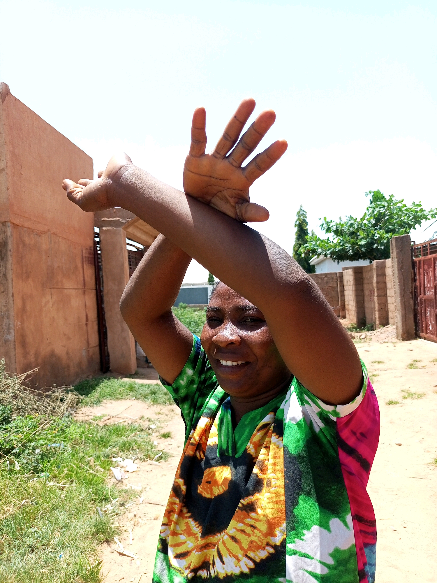 A medium shot captures a smiling woman with dark skin and short hair, wearing a brightly colored tie-dye shirt, in an outdoor setting. Her arms are raised above her head, with her hands open and fingers spread, as if signaling or dancing. The background suggests a dry, possibly rural environment with a dirt path, low green vegetation, and earthen-colored buildings with some fencing visible. The sky is clear and bright. The location is identified as Tempure, Nigeria.