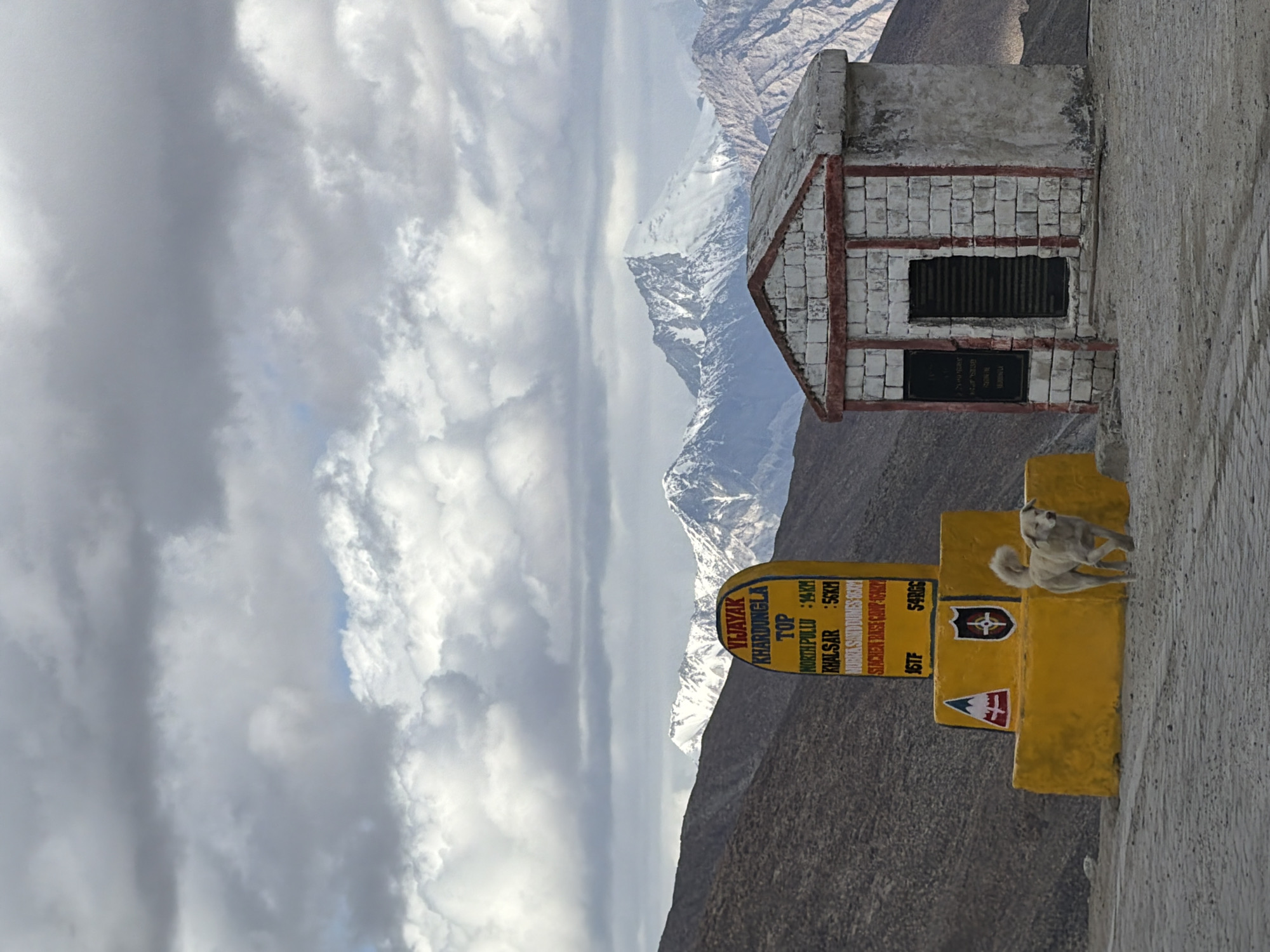 An overcast sky looms above a rocky, mountainous landscape. In the foreground, a yellow directional signpost, adorned with military insignias, stands next to a small, stone structure. The signpost indicates distances to various locations, including "KHARDUNG LA TOP," "NORTH PULLU : 14KM," "KHALSAR : 56KM," and "HUNDER SAND DUNES 56KM." A white dog trots past the signpost. The structure to the right is a rudimentary building with a slatted door and a dark rectangular panel on the left side. The background is dominated by snow-capped mountains. This scene appears to be located at Khardung La Pass, a high mountain pass in the Ladakh region near Leh, India, which is known for its stunning mountain vistas and its status as one of the world's highest motorable passes.