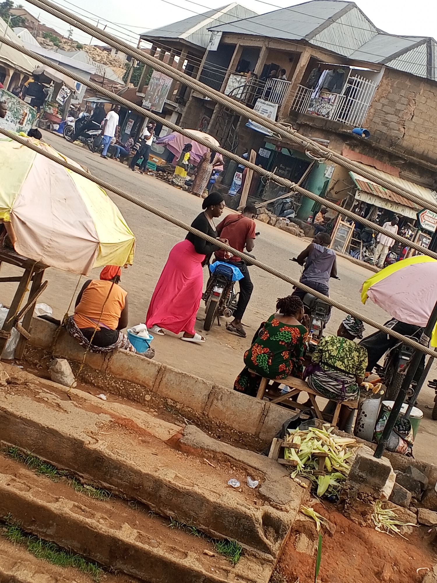 The image depicts a street scene in Gwong, Nigeria. A woman in a bright pink skirt walks past a woman sitting on a low wall next to a yellow and white umbrella. Two men on motorcycles are stopped in the street nearby. Further down the street, people walk along the sidewalk and stand in front of shops. Several buildings with balconies and awnings line the street. Overhead, thick cables crisscross the frame. Corn husks are gathered on the ground near a group of women sitting on a bench. Steps lead up to the area where the woman in the orange shirt is sitting.