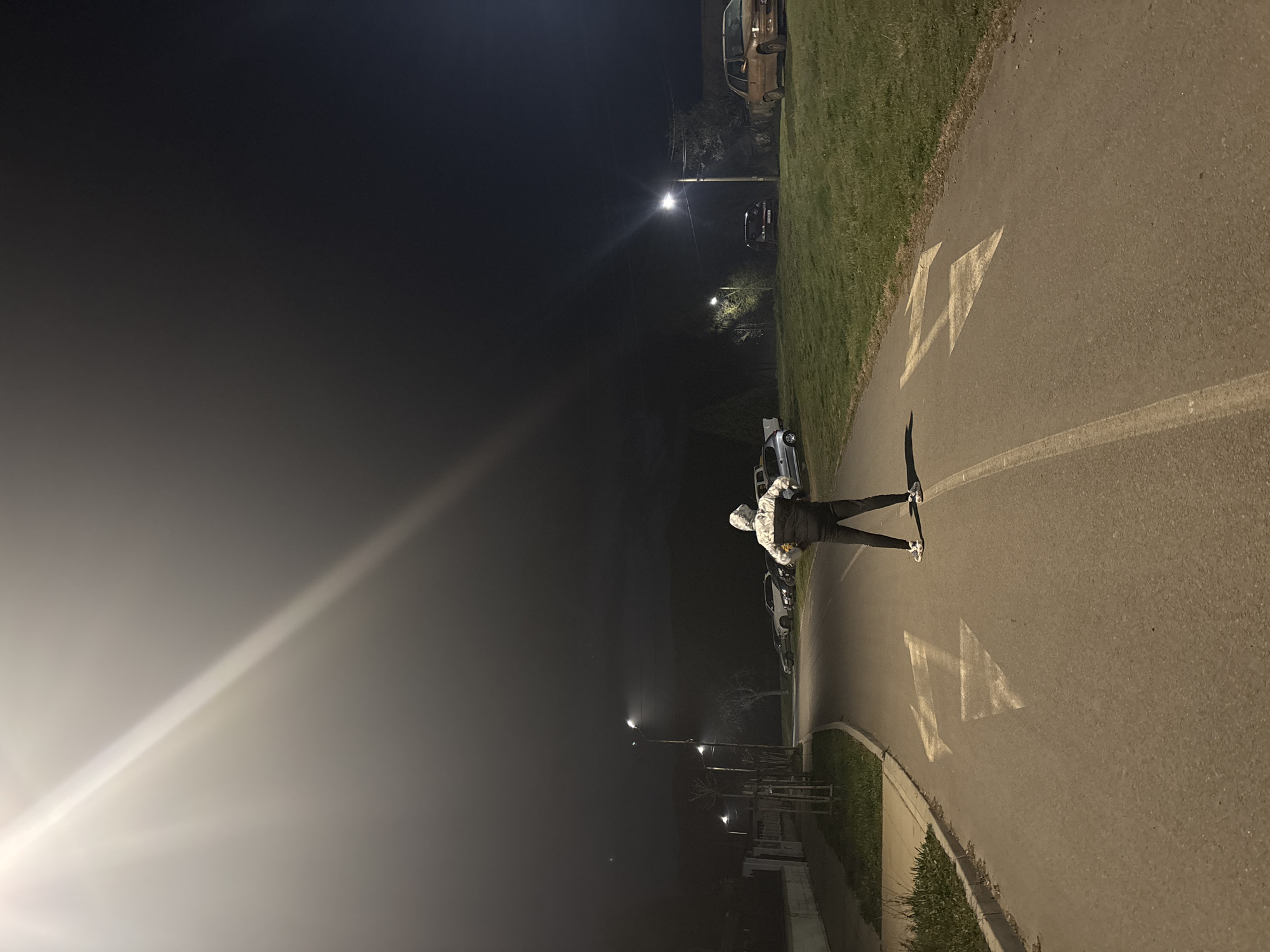 A nighttime scene in Zlatitsa, Bulgaria, shows a person standing in the middle of a paved road marked with directional arrows. The road is flanked by a grassy area on one side and a sidewalk or curb on the other. Streetlights illuminate the road and surrounding area. Several parked cars are visible along the side of the road. The sky is dark, with a foggy or hazy quality. The person is wearing dark pants, white shoes, and a light-colored hooded jacket.