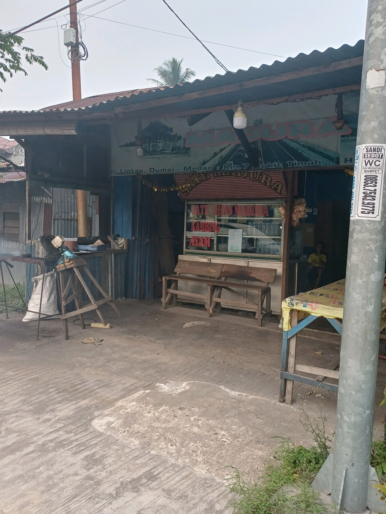 The image depicts a modest outdoor food stall with a corrugated metal roof, situated on a paved area. The stall has a display window showcasing food items and a wooden bench in front. In the background, a person is seated inside the stall. A utility pole with electrical wires is visible on the right side of the image, and a sign with Indonesian text is attached to it. The scene appears to be in an urban or semi-urban environment, potentially in Dumai City, Indonesia, given the context. The overall impression is that of a simple, functional establishment.