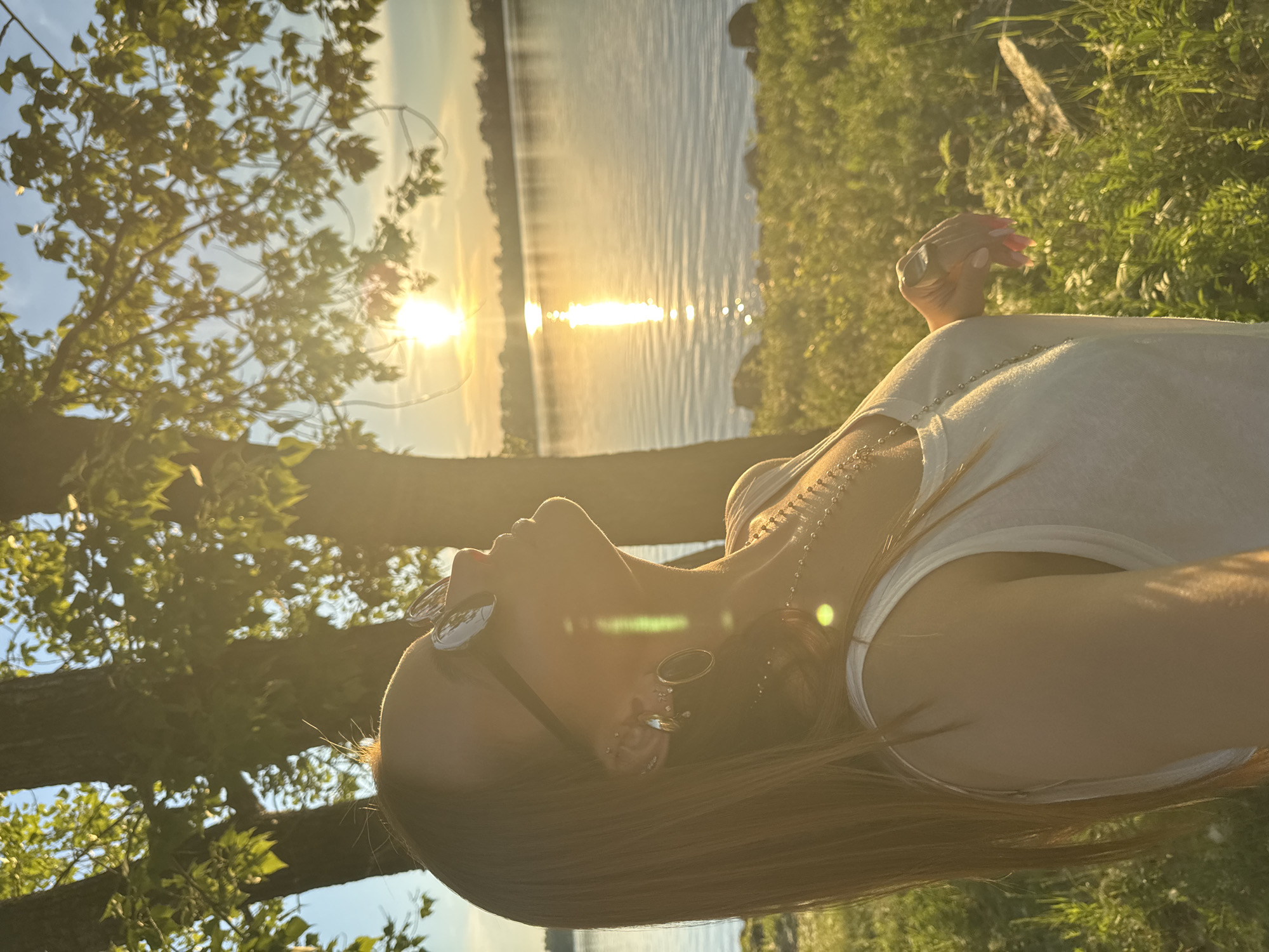 A woman with long, auburn hair, wearing a white tank top and sunglasses, is captured in a profile view looking upwards towards the setting sun. She is adorned with multiple earrings, including large hoop earrings, and several necklaces. The scene unfolds outdoors, with a body of water, likely a river or lake, in the background. The sun is low on the horizon, casting a warm, golden light and creating a reflection on the water. Trees with green foliage frame the top portion of the image, and lush green grass covers the foreground and mid-ground. The location is specified as the city of Chaikovsky, Russia.