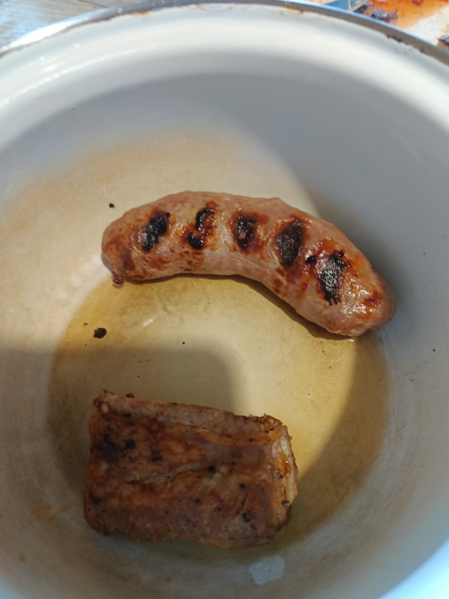 The image shows two pieces of cooked meat, a sausage and what appears to be a strip of bacon, in a white pot or pan. The sausage is curved and has grill marks. The bacon is rectangular and appears to be well-done. There are dark spots and some grease visible in the pot. The background is blurred but appears to be a surface, possibly a table. The image is a close-up of the food. There is no indication of the city of Varna, Bulgaria in the image.