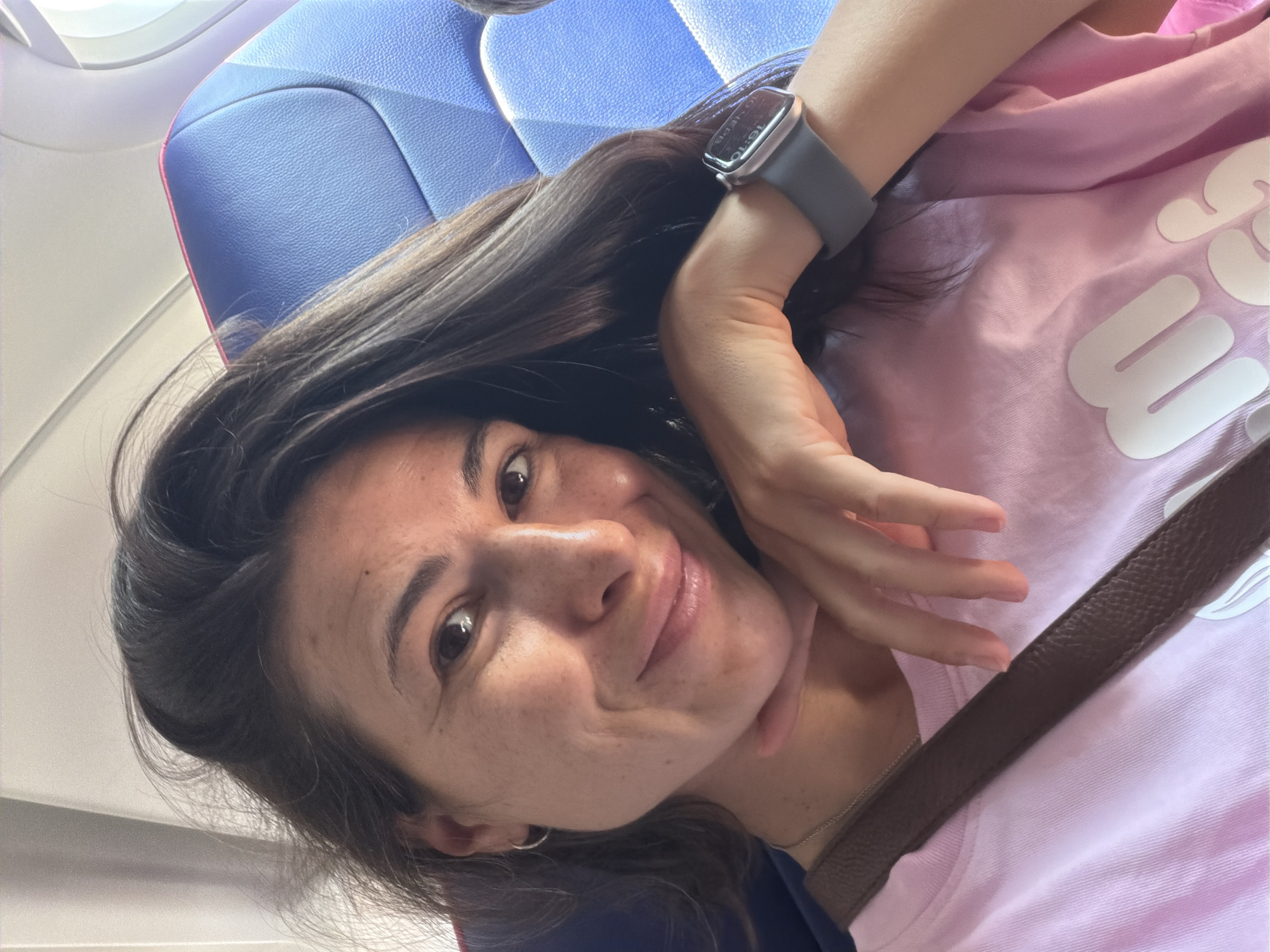A woman with long, dark hair is taking a selfie in an airplane seat. She is wearing a pink t-shirt and has a brown strap across her chest. She is smiling and resting her chin on her hand. The airplane seat is blue. The image does not provide any information about the city of Donja Cijevna, Montenegro.