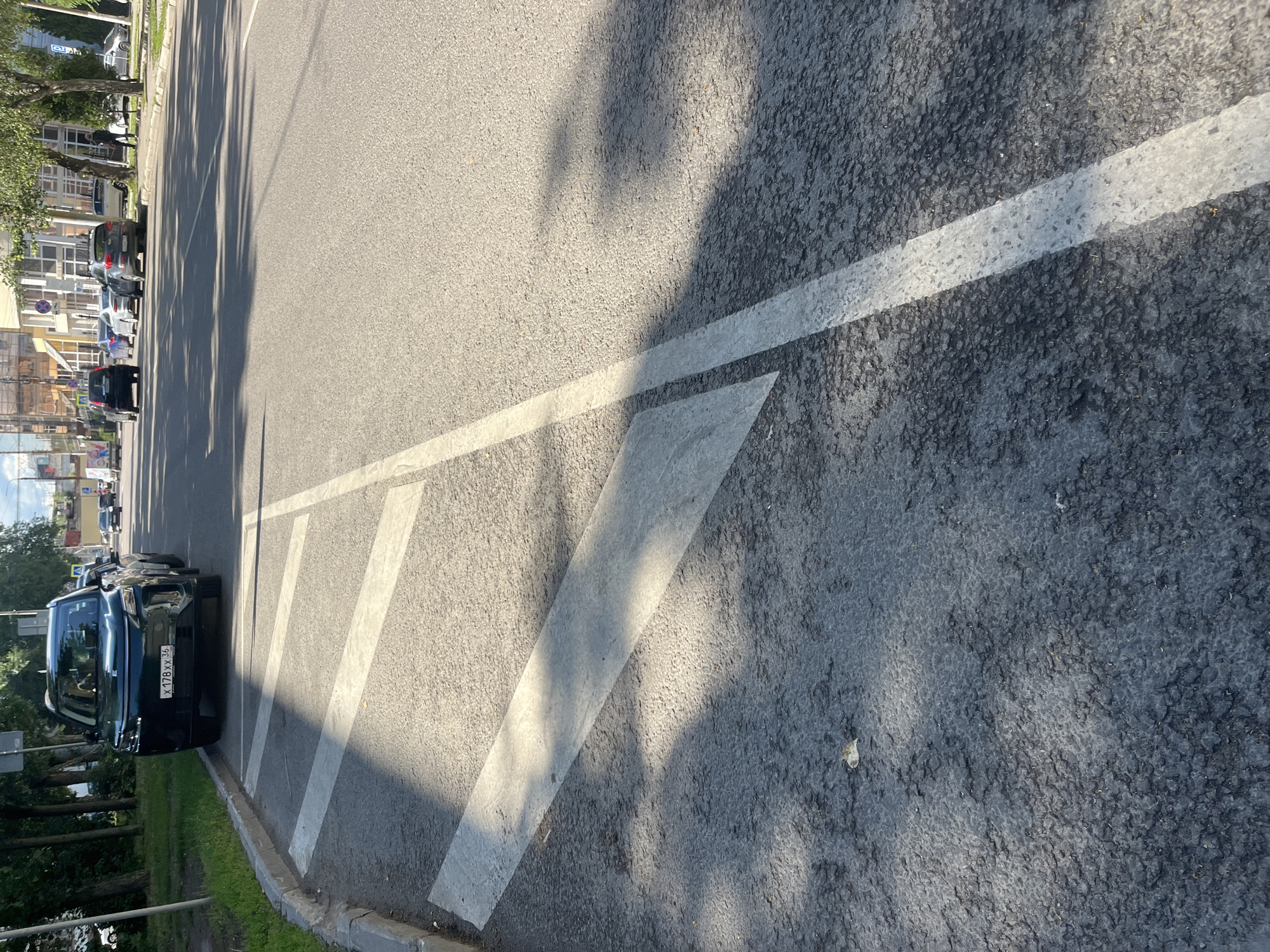 The image captures a street scene in Novaya Usman, Russia, bathed in the bright sunlight of what appears to be daytime. The perspective is from ground level, looking down a paved road marked with white pedestrian crossing lines. The asphalt is a mix of dark grey and lighter patches, with visible texture. The white lines are broad and clearly defined, with some appearing more worn than others.
On the left side of the road, a sidewalk runs alongside, bordered by a concrete curb. Beyond the sidewalk, a line of trees with green foliage creates a dappled shadow pattern on the road and sidewalk. Behind the trees, buildings are visible, featuring multiple windows and a light-colored facade, suggesting an urban or suburban setting. Cars are parked along the side of the road and some are driving by. One black car in the foreground is particularly noticeable, parked facing the right edge of the frame. Its license plate, with the visible text "X 178XX 36", is legible.
The overall scene depicts a typical day on a street, with the natural elements of sunlight and shadows playing a significant role in the visual composition. There are no people prominently featured in the immediate foreground, but they are implied by the presence of the pedestrian crossing and the parked cars. The atmosphere seems calm and ordinary, typical of a sunny day in a settled area.