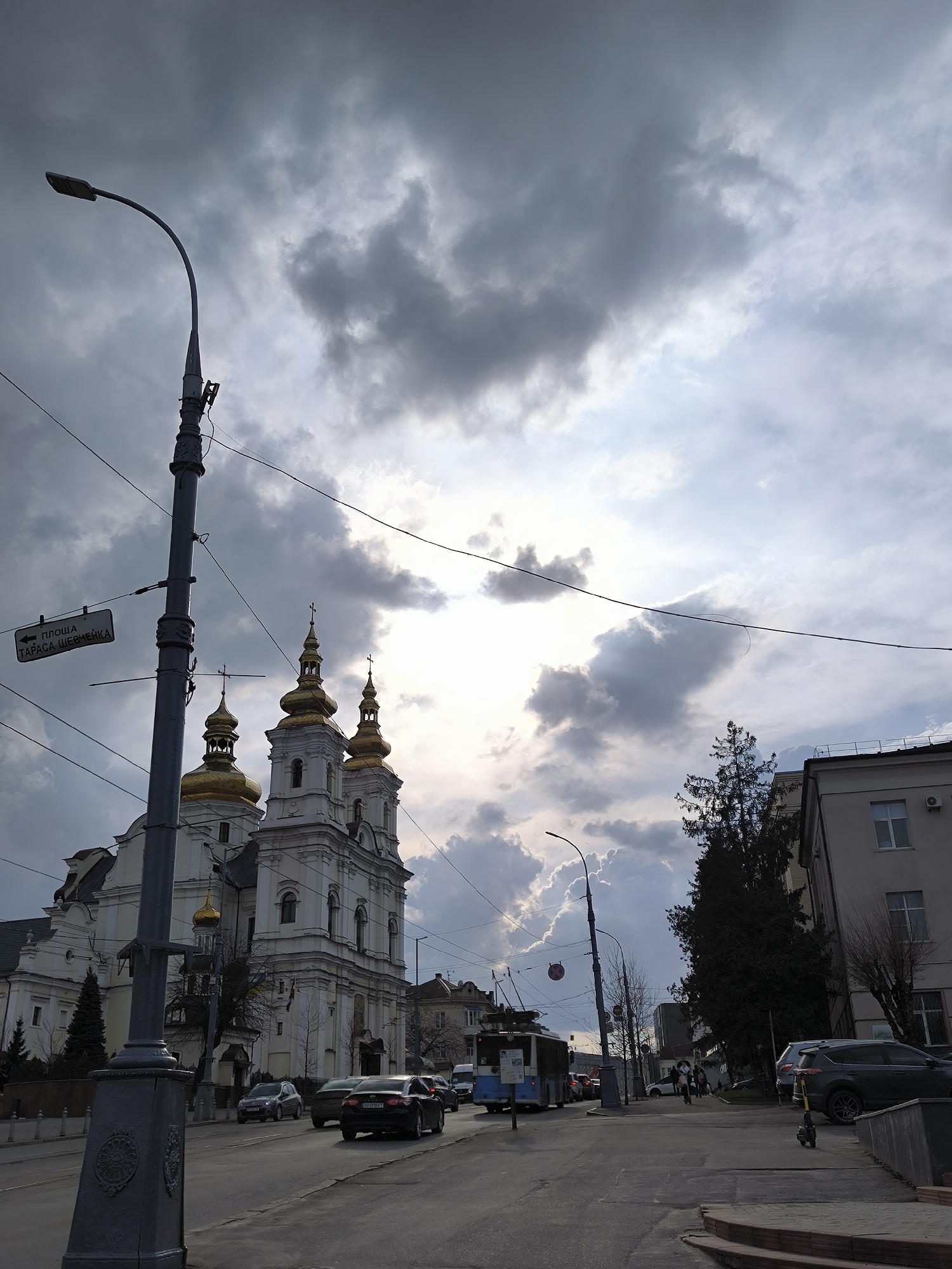 The image shows a street scene in Vinnytsia, Ukraine. In the foreground is a street with cars and a trolleybus. A pedestrian is crossing the street. A street lamp with a sign pointing towards "Ploscha Tarasa Shevchenka" (Taras Shevchenko Square) is on the left. The white and gold-domed Cathedral of the Holy Transfiguration is on the left side of the street. Buildings line the right side of the street. The sky is overcast with clouds.
