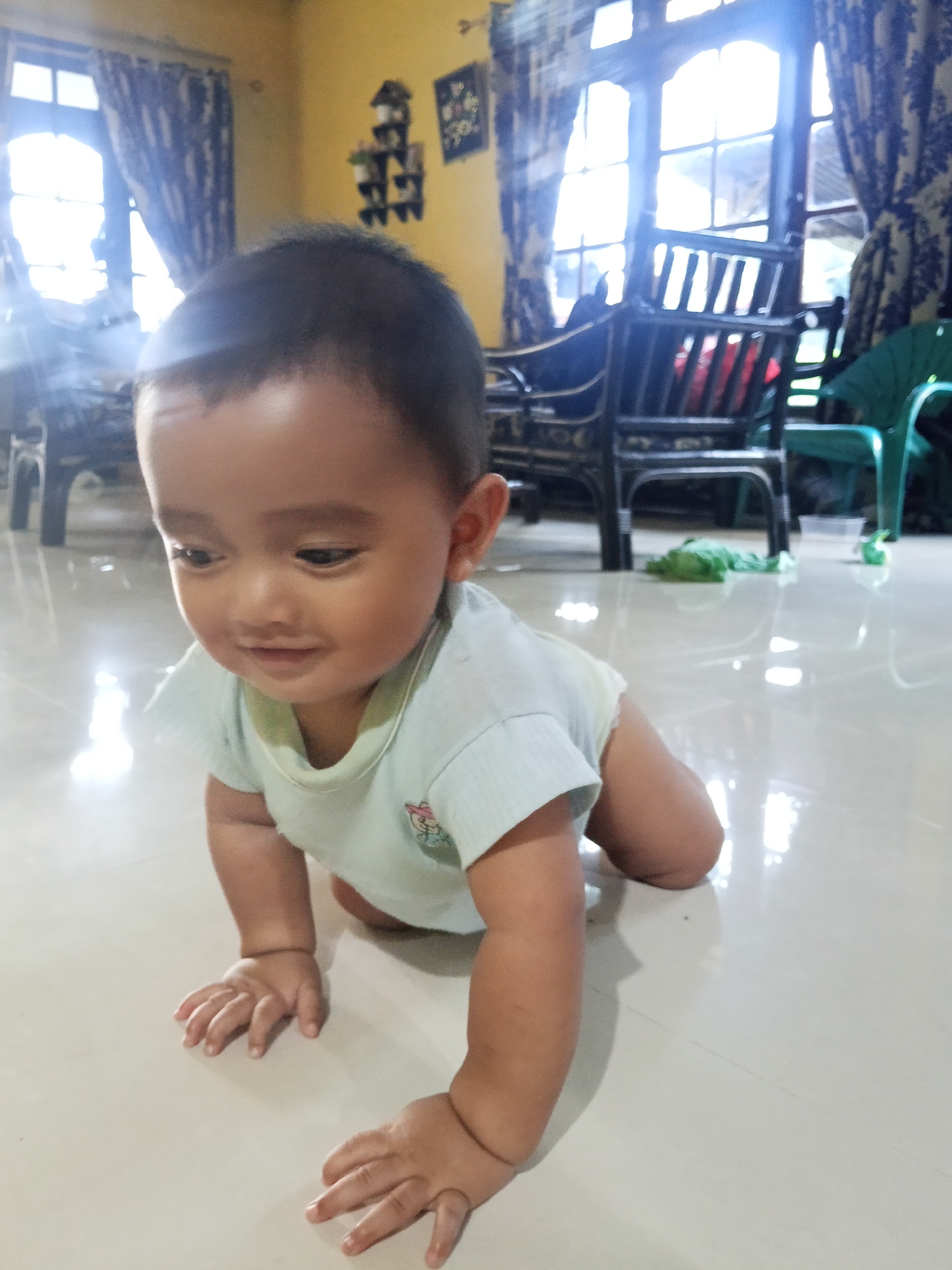 A young child is in a crawling position on a polished floor. The child is wearing a light blue onesie. The room appears to be a living area, with large windows covered by patterned curtains, a dark-colored chair with woven details, and another green chair in the background. The floor reflects the light from the windows. The scene is indoors, likely within a residential setting in Lestari Indah, Indonesia.