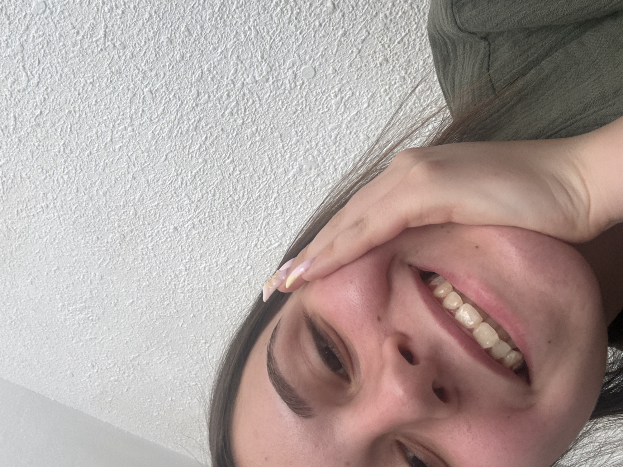 The image is a close-up, low-angle selfie of a young woman's face and upper body. She has dark hair and is wearing a green top. Her right hand is touching her cheek, and she is smiling, showing her teeth. Her fingernails are long and appear to be painted with a light, multi-colored design. The background is a white, textured wall. The image does not contain any information about the location in Burgas, Bulgaria.