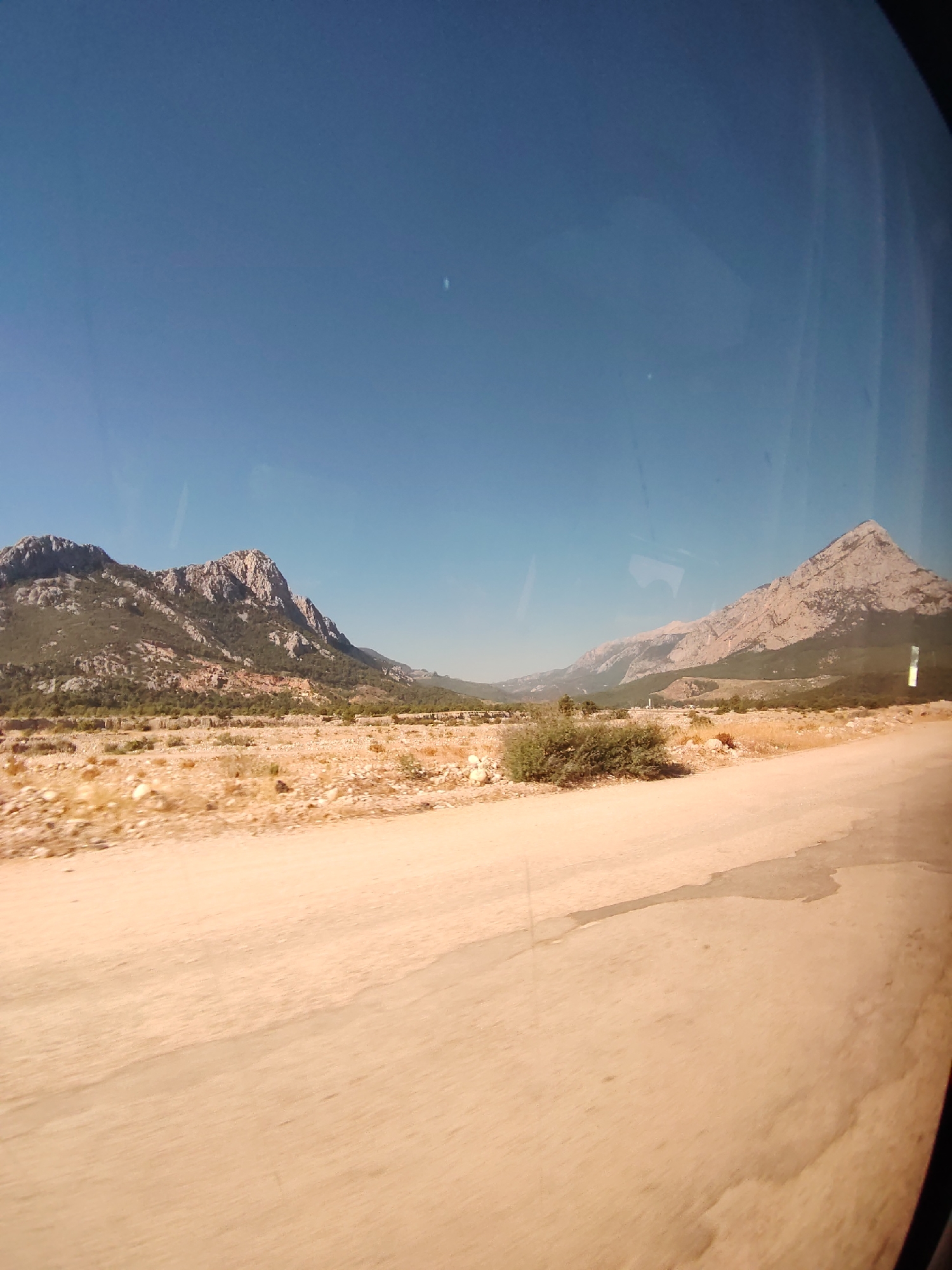 The image shows a daytime outdoor scene, likely taken from a vehicle moving along a dusty, unpaved road. The foreground is dominated by the beige, textured surface of the road, with some patches of dry, sparse vegetation and scattered rocks. In the midground, a flat, arid landscape stretches towards a range of mountains. These mountains are rugged and rocky, with slopes covered in sparse, green vegetation and bare, exposed rock faces. The sky is a clear, deep blue with no visible clouds. The scene suggests a rural or semi-arid environment, consistent with the natural landscapes found around Konyaaltı, Turkey, which is known for its mountainous terrain and proximity to the Mediterranean coast. No people or specific man-made structures are clearly identifiable within the frame, beyond what might be implied by the road itself. The lighting indicates bright daylight, casting shadows that are not strongly defined due to the overcast haze or the angle of the sun.