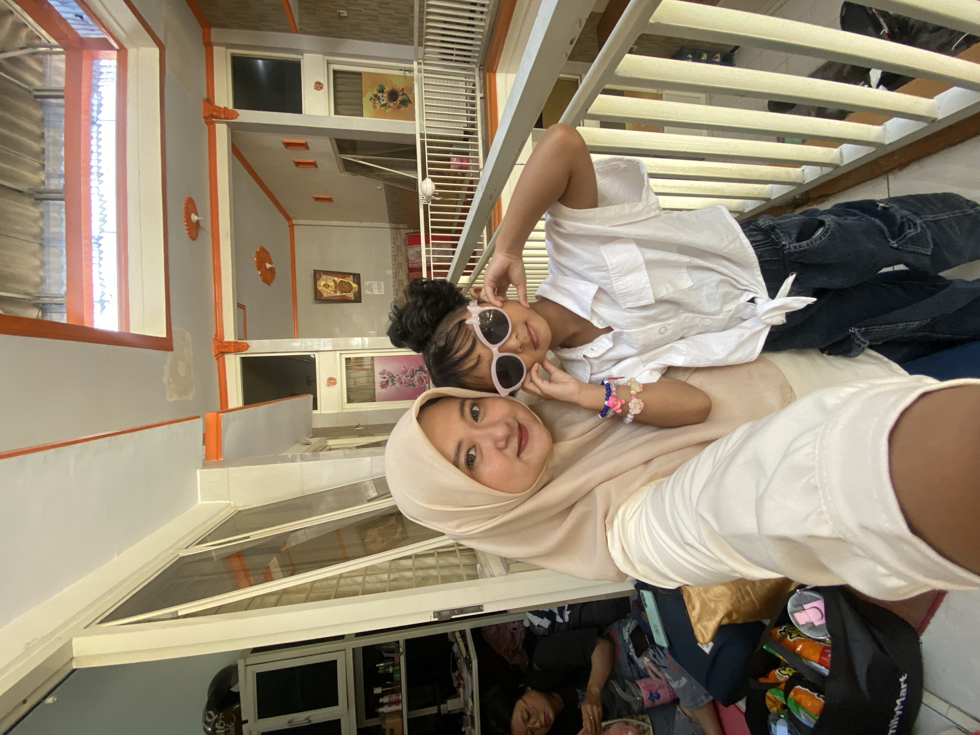 Two individuals are captured in a selfie. The person in the foreground wears a light-colored hijab and a matching light-colored shirt or dress. A child in a white shirt and dark jeans is positioned behind her, wearing sunglasses. They are lying on a balcony behind a white railing, with a cluttered room visible below through a window. A FamilyMart bag is in the foreground. The location appears to be a residence in Jember, Indonesia, evidenced by the architectural style and visible products.