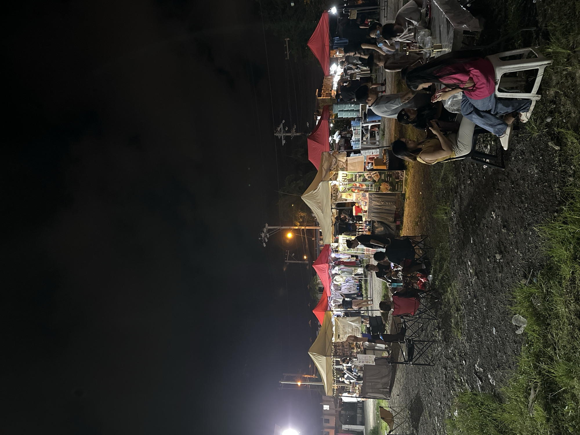 The scene depicts a night market in Davao City, Philippines, conducted outdoors at night under a dark, possibly overcast sky. The ground consists of uneven, dark earth mixed with gravel and sporadic patches of green grass, indicative of a temporary or undeveloped lot. Illumination is provided by overhead string lights within the market area, supplemented by distant streetlights, one prominent on the far left, and another further back near utility poles and overhead power lines.
The market features multiple pop-up canopy tents of varying colors, including beige/tan and red, sheltering vendor stalls. Vendors attend to stalls displaying various goods, including clothing racks under a red tent and packaged food items, bottles, and jars under other tents. A visible menu board details food items such as "MUSUBI," "CHICKEN," and "CHEESY," while another sign identifies "Aree Coffee." Tables, some covered with white tablecloths, and an assortment of folding chairs and plastic stools are arranged for customer use.
Numerous individuals, estimated at 20-30, populate the scene. In the foreground to the right, two adult females are seated, both engaged with mobile phones. One wears a yellow top and light trousers, sitting on a black folding chair; the other wears a pink long-sleeved shirt and blue jeans, seated on a white plastic stool. Towards the center-left, a group of approximately three to four males are seated around a small table with folding chairs, appearing to dine and converse. Other individuals are observed standing, browsing stalls, or moving through the market space. In the background, dimly lit residential structures and foliage are discernible, suggesting an urban or suburban setting.