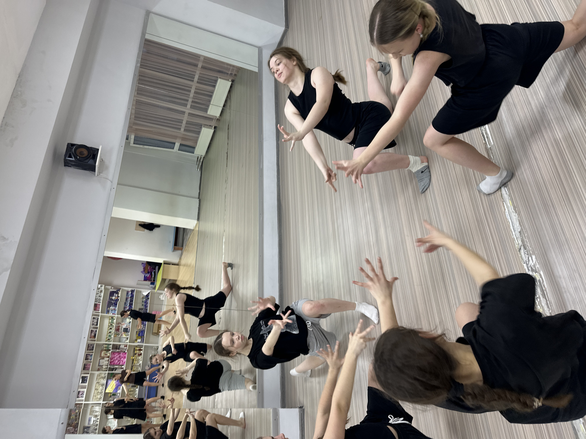 The image captures an active dance practice session in an indoor studio in Votkinsk, Russia. Several young females, appearing to be teenagers or young adults, are engaged in various dance or movement exercises. They are dressed in athletic attire, primarily black tank tops, t-shirts, and shorts, with some wearing grey shorts or white/grey socks. Their poses are dynamic, featuring outstretched arms, expressive hands, bent legs, and focused expressions, suggesting a choreographed routine or warm-up.
A prominent feature of the setting is a large mirrored wall that spans much of the room, reflecting the dancers and the opposite side of the studio. This creates a sense of depth and doubles the visible number of participants. In the foreground, one dancer is seen from behind with her long brown hair braided, while others are caught mid-motion or in kneeling positions.
The flooring is made of light-colored wood or laminate. On a white wall adjacent to the mirrored surface, a black speaker is mounted, indicating that music is used during practices. The reflections in the mirror reveal more details of the room, including shelves filled with numerous trophies, awards, and what appear to be framed photos or certificates. These items suggest that the location is an established dance school or studio with a history of achievements.
The lighting within the studio is bright and uniform, typical of an indoor practice space. The time of day and external weather conditions are not discernible from the image. No visible text is clearly legible anywhere in the photograph. The overall scene conveys a dedicated and energetic environment for dance training.