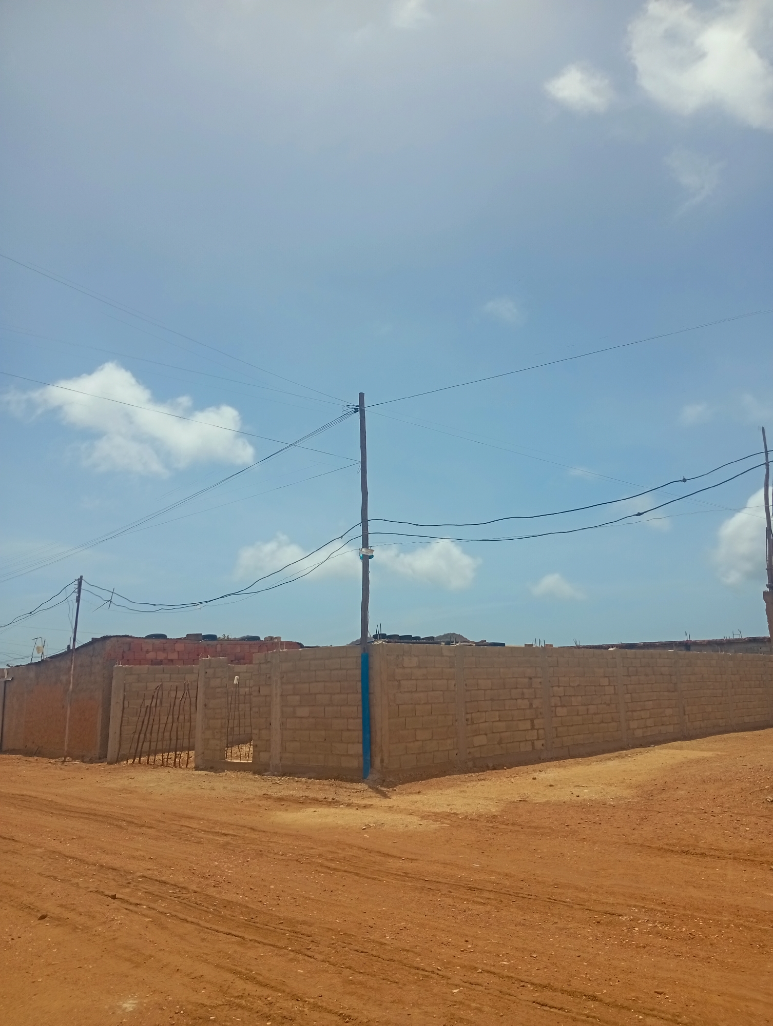 The image depicts a construction site in Altagracia, Venezuela, under a bright, partly cloudy sky. A tall, rough-hewn wooden pole stands vertically on the right side of the frame, supporting multiple power lines that stretch horizontally across the sky. To the left of this pole, another similar pole is visible, also supporting power lines.
In the foreground, a dirt road with tire tracks is prominent, suggesting vehicular activity. A low, unpainted wall constructed from concrete blocks forms a barrier across the middle ground of the image. Part of this wall appears to be under construction, with exposed rebar and unfinished brickwork visible in sections. Some discarded tires are stacked on top of a section of the wall. The wall turns a corner, extending to the right edge of the frame.
On the far left of the image, another section of wall, possibly older or made of a different material, is visible. This section appears to be made of compacted earth or stucco. A gate-like structure made of vertical wooden poles is set into an opening in the concrete block wall. The overall impression is one of ongoing construction or development in a semi-arid environment.