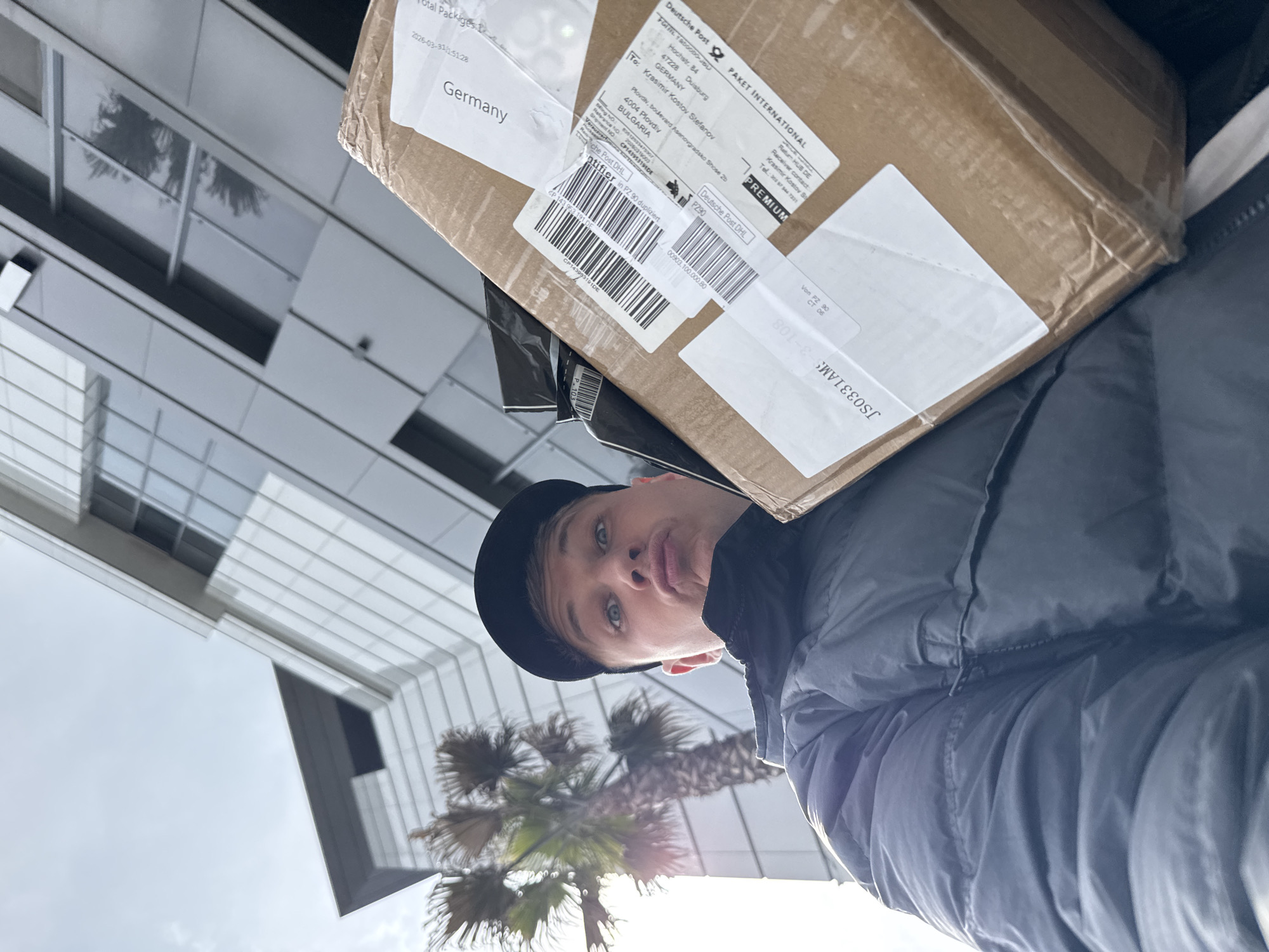 A man is holding a brown cardboard box with German postage labels in front of a modern building and a palm tree. The box is addressed to Krasimir Kostov Stefanov in Plovdiv, Bulgaria, specifically boulevard Asenovgradsko Shose 2b. The sender is Deutsche Post, from Duisburg, Germany. The man is wearing a black cap and a dark jacket. The building has a modern architectural style with reflective glass panels.