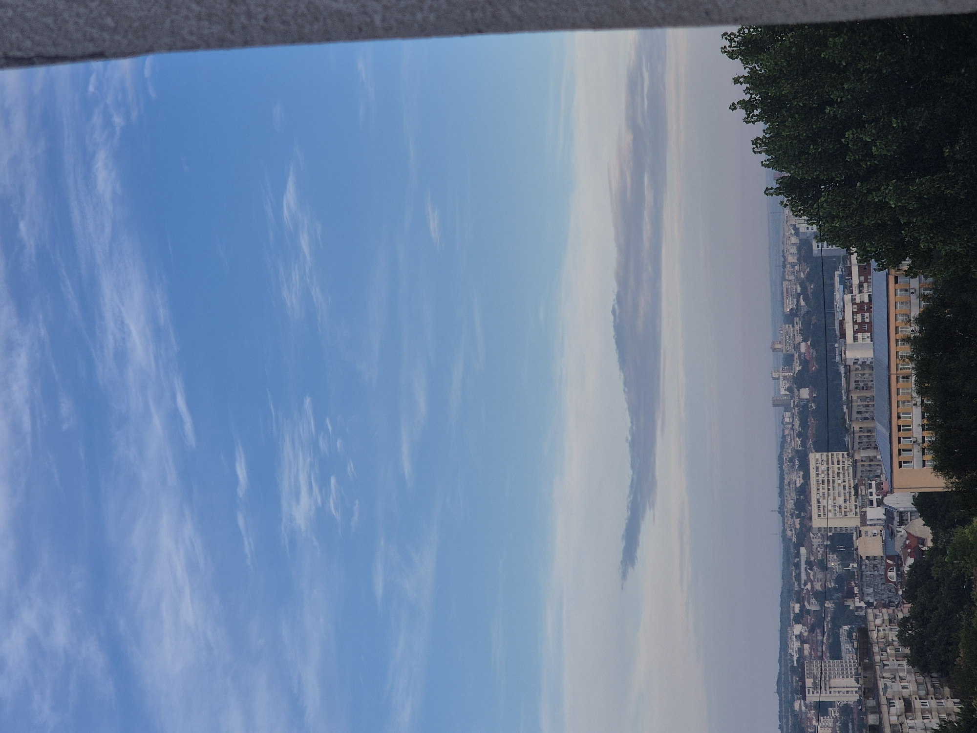 An elevated, wide shot captures a cityscape under a bright blue sky dotted with wispy white clouds. The image is framed on the right by a textured, light-gray vertical element, possibly a wall or pillar, which partially obscures the view. The city itself is a dense collection of buildings of varying heights and architectural styles, with residential blocks, some taller apartment buildings, and commercial structures evident. A prominent yellow and orange building stands out in the foreground, partially obscured by dark green trees. The urban sprawl extends towards a hazy horizon, where more distant buildings and possibly a body of water are faintly visible. The overall impression is of a daytime scene in an urban environment.