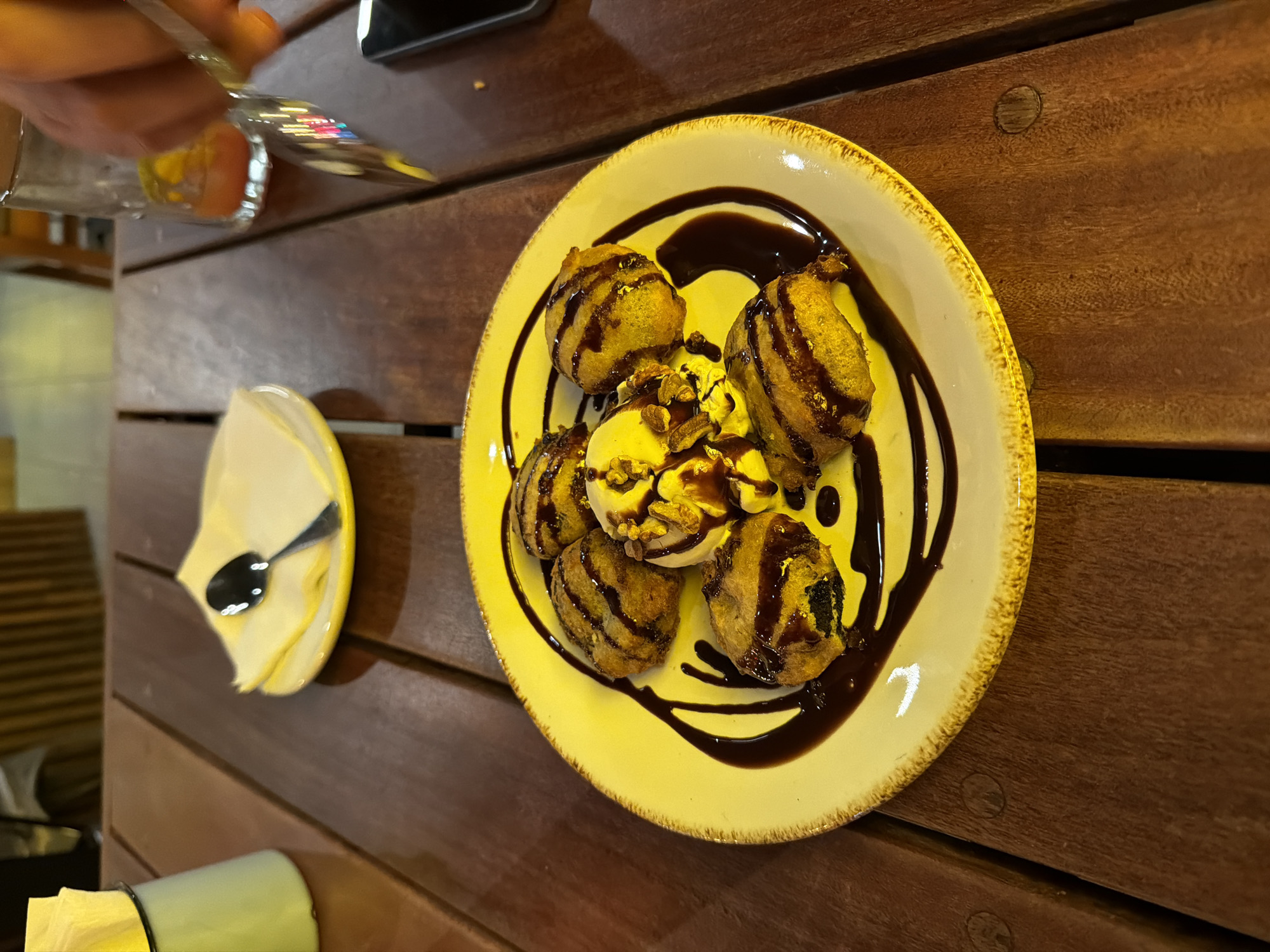 A wooden table holds a plate of fried ice cream drizzled with chocolate sauce. Beside the plate is a smaller dish with a spoon. A hand holding silverware is visible in the upper left corner. The location is a restaurant in an unknown city.
