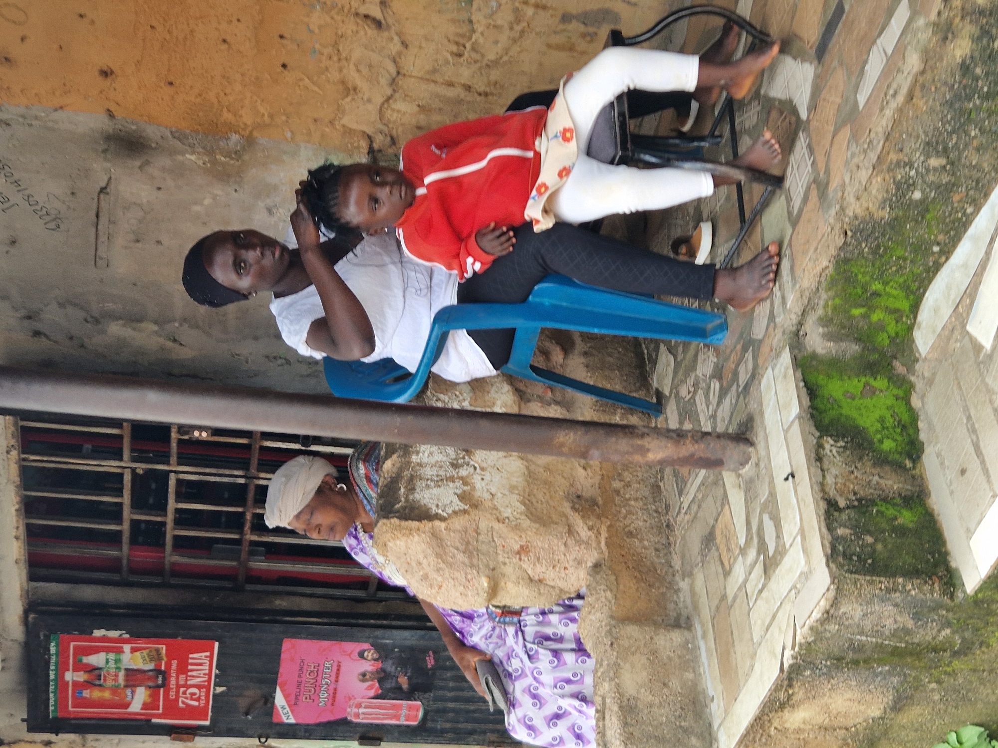 In a street scene of an unknown city, a woman with a dark headscarf is braiding the hair of a young girl. The woman is seated on a blue plastic chair, wearing a white top and black leggings, and is focused on her task. The girl, sitting on a smaller stool, is wearing a red zip-up jacket over a light-colored patterned skirt and white leggings. Beside them, an older woman in a purple and white patterned dress and a white headwrap sits on a raised concrete ledge, holding a grey clutch. Behind the older woman, posters are visible on a storefront, one featuring beverages and another an energy drink. The environment appears to be an outdoor area with a concrete floor tiled with various stone patterns, and moss growing on a weathered concrete wall. A thick metal pole stands vertically in the foreground.
