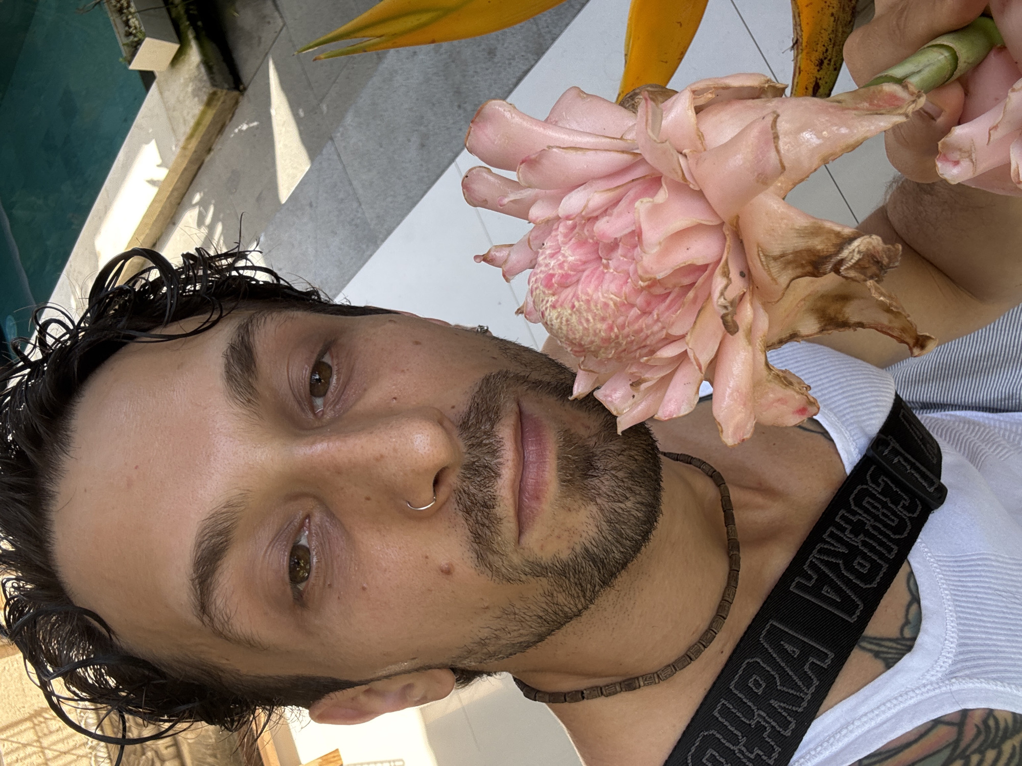 The media file is a close-up, vertical shot of a man taken outdoors in a tropical setting, possibly a selfie. The man, with dark, curly, wet hair and stubble facial hair, looks directly at the viewer with light brown eyes. He has a silver nose ring and is wearing a white tank top and a dark, beaded wooden necklace. A tattoo is visible on his left arm or shoulder.
He is holding a large, pink torch ginger flower (Etlingera elatior) in his right hand, which is prominently featured in the upper right quadrant of the image. The flower shows signs of natural browning and wear on its petals.
The background indicates a bright, sunny day with strong shadows on light-colored paving. A turquoise swimming pool is visible to the left, and lush green foliage, along with distinctive yellow and orange bird of paradise leaves, are present in the upper background, confirming a tropical environment consistent with Ubud District, Indonesia.
The man's expression is neutral. A black strap, likely from a bag or camera, crosses his chest. Partial white text, "RA I JCORA", is visible on this strap.