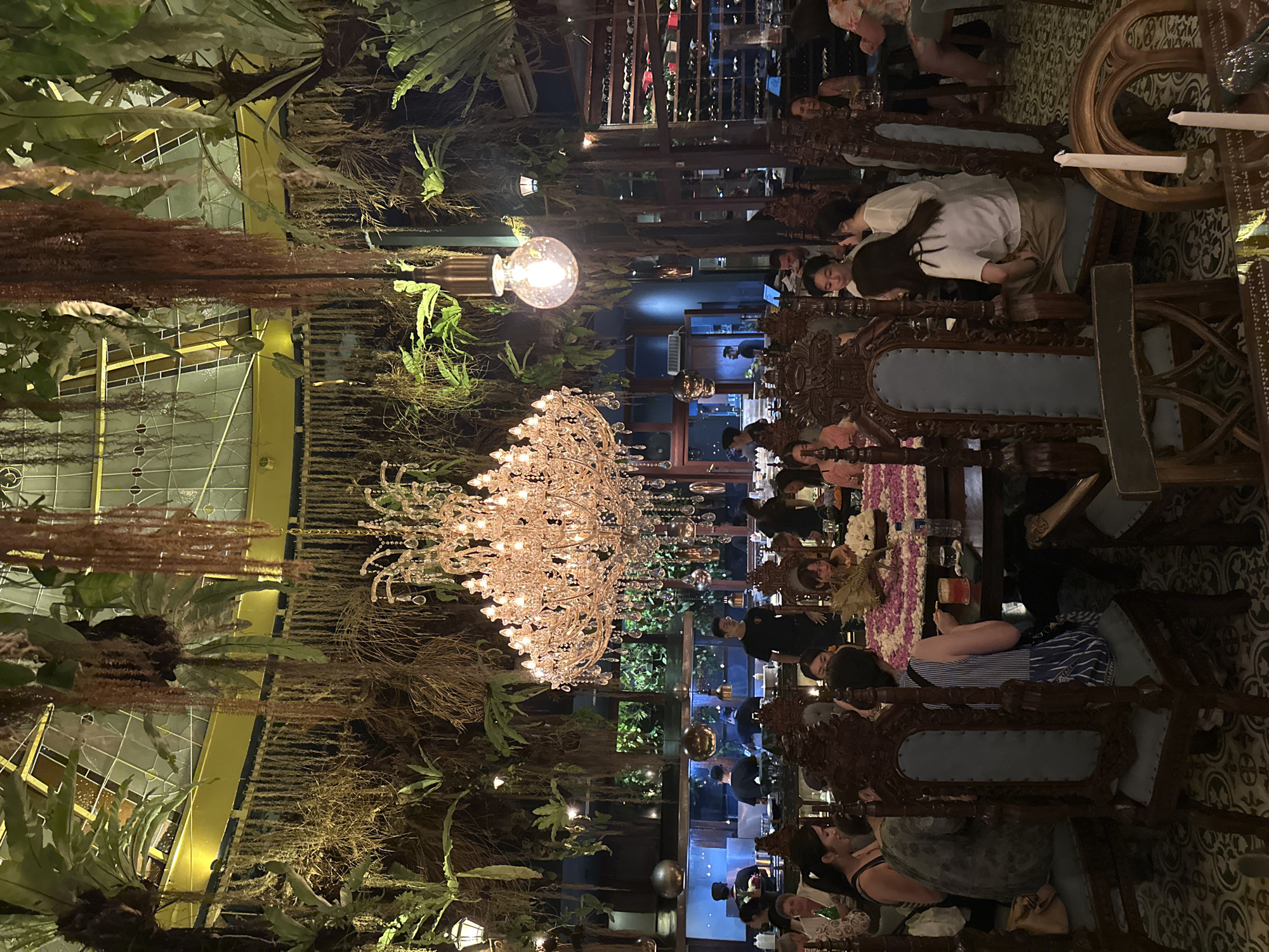 The image depicts the interior of a restaurant in Ubud District, Indonesia. The setting suggests a high-end dining experience, with ornate wooden chairs, elaborately set tables, and crystal chandeliers. Numerous diners are seated at the tables, engaged in conversation. The restaurant's decor includes extensive greenery, with plants and vines cascading from the ceiling, blending natural elements with the formal dining atmosphere. Other light fixtures such as glass bulbs are visible. In the background, a wine rack can be seen. Staff members in black uniforms are present. The scene has a bustling yet elegant ambiance.