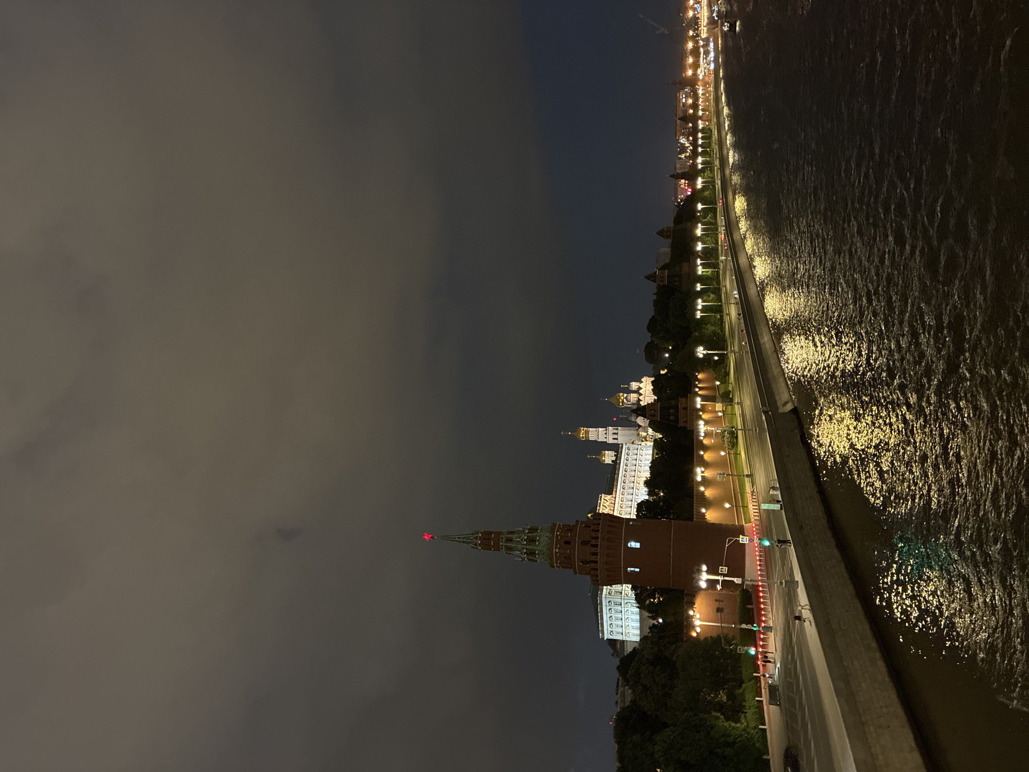 A nighttime view of the Moscow Kremlin complex in Moscow, Russia, is positioned alongside a river. Prominent buildings include a red brick tower topped with a red star, other white and gold domed structures within the complex, and adjacent illuminated buildings. A road with streetlights and moving traffic runs along the edge of the Kremlin. A river reflects light with dark ripples along its surface. The sky is predominantly dark with some cloud cover.