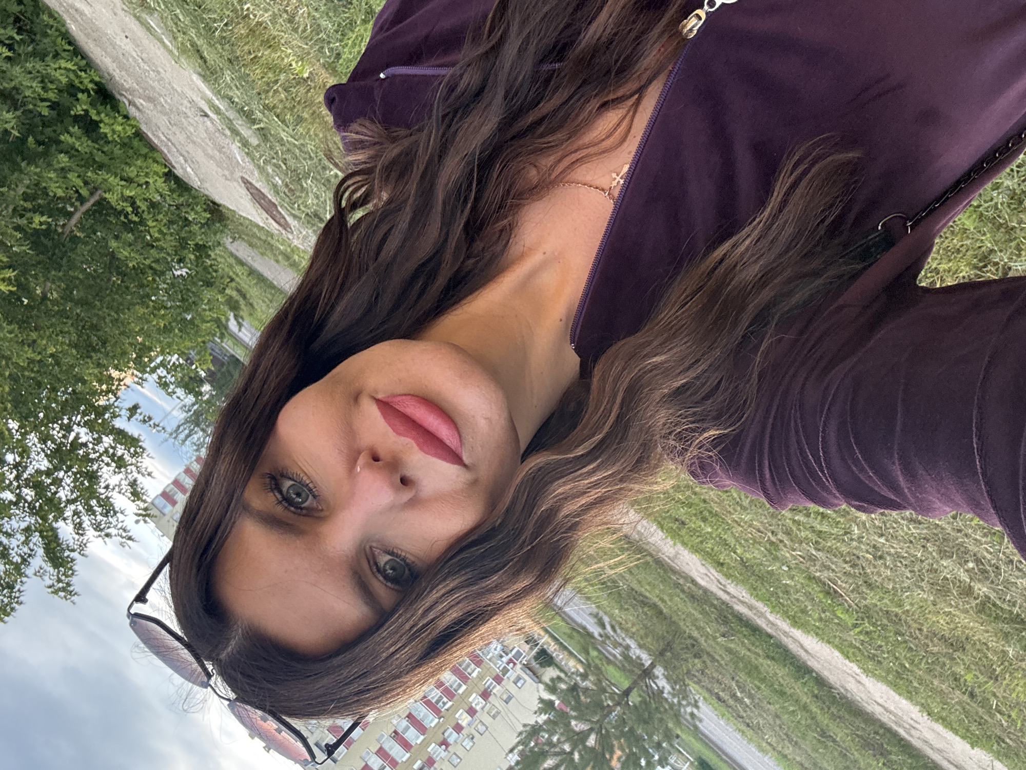 A young woman is taking a selfie outdoors, holding the camera above her head and angling it down towards herself. She is wearing a dark purple zippered jacket, a delicate necklace with a small cross pendant, and sunglasses perched on her head. Her long, dark brown hair is mostly loose, with some strands framing her face. She has on light pink lipstick and appears to be looking directly at the camera with her green eyes.
The background shows a slightly overcast sky with some greenery and trees on the left side. On the right, there is a pathway or dirt road running through a grassy area. In the distance, a multi-story apartment building with a light-colored facade and red window frames is visible. The overall impression is that of a casual outdoor setting, possibly in a park or residential area. The lighting suggests it is daytime, likely late afternoon or early evening given the angle of the light. There is no visible text in the image. The woman's expression is neutral to pleasant.