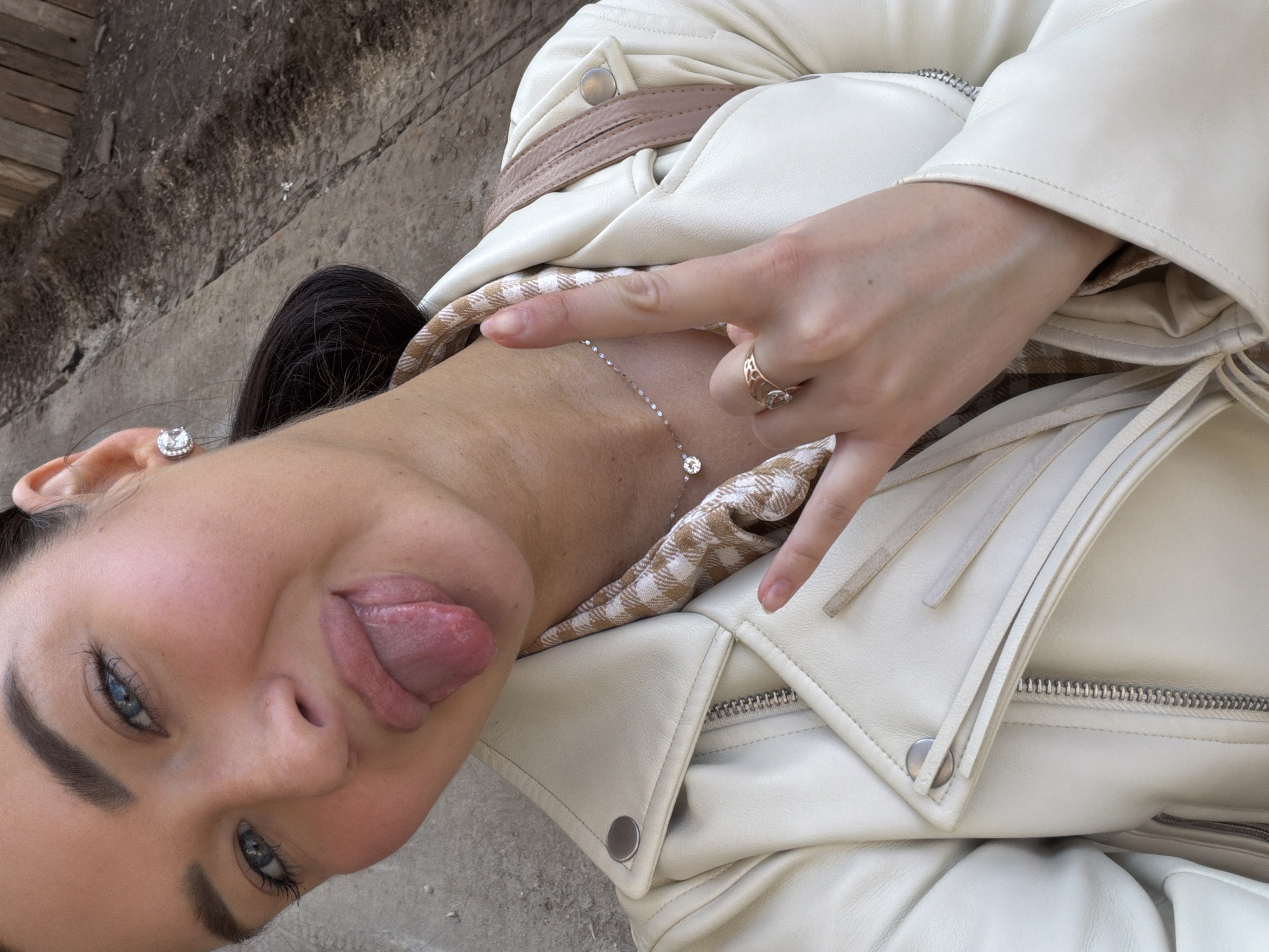 A young woman, wearing a white leather jacket, is sticking her tongue out towards the camera. She is wearing a silver necklace with a pendant, and a ring on her right ring finger. The photo appears to have been taken in an urban environment, with a textured concrete surface visible in the background. I cannot determine if the photo was taken in Moscow, Russia.
