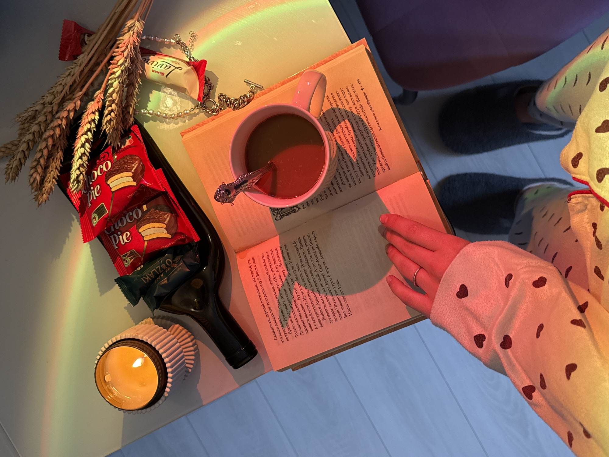 An overhead, slightly angled photograph depicts an indoor tabletop scene in Unknown, Unknown. A light-colored surface, presumably a table, holds various items. On the left, a lit candle in a white, fluted holder casts a warm glow. Adjacent to it lies a dark glass bottle, horizontally oriented. Stacked on and around the bottle are packaged snacks, including two red "Choco Pie" packages, a partially visible green package, and a cream-colored "Ülker Laviva" bar. A small bundle of dried wheat stalks is positioned behind the snacks.
At the center, an open book with Cyrillic script text is displayed, with page 159 visible on the right page. A light pink ceramic mug containing a dark liquid with a silver spoon rests on the book. Next to the book and mug, a silver-colored chain necklace with a T-bar clasp and a strand of pearls are visible.
A person's left hand, adorned with a simple ring on the ring finger, is resting on the open book. Below the table, the person's lower body is visible, wearing a light-colored garment with dark heart patterns on the sleeve and light-colored patterned loungewear. Dark, soft slippers are on the light-colored wooden or laminate floor. A portion of a dark purple chair is visible on the right side of the frame.
A prominent rainbow light spectrum is projected across the entire scene, originating from the left and sweeping across the objects, illuminating them with distinct bands of color from blue to red.