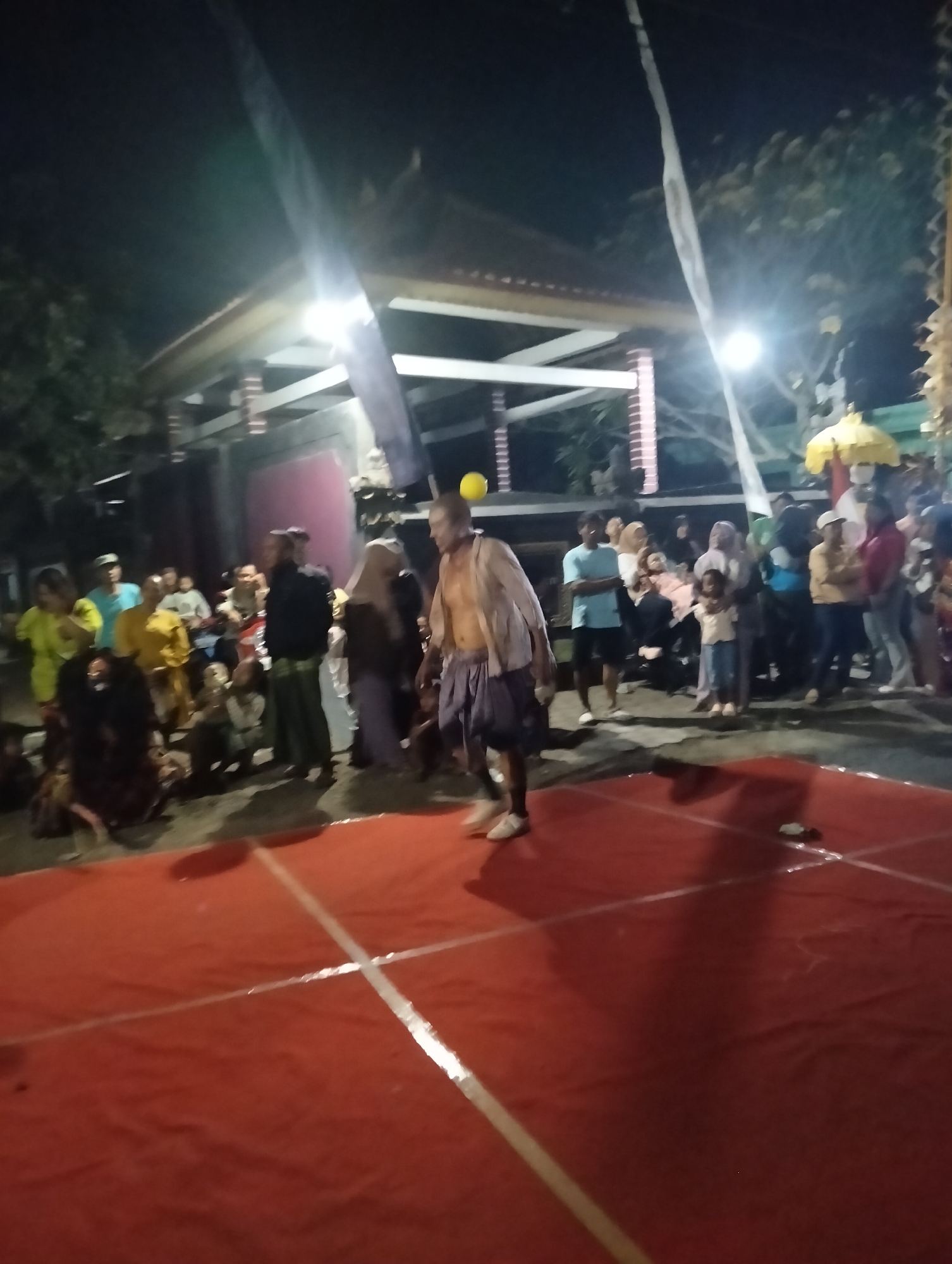In Sumbersewu, Indonesia, at night, a performance takes place on a red carpeted stage marked with white tape. A shirtless man with a yellow object on his head, wearing dark shorts, is the central performer. He stands facing a crowd gathered on the dark ground behind the stage. The crowd, composed of men, women, and children, are dressed in casual attire, with some women wearing headscarves. Behind the crowd, a structure with a pitched roof and supporting columns is visible, illuminated by overhead lights. Trees and foliage surround the area, with a yellow umbrella-like decoration visible on the right side of the frame. The overall atmosphere suggests a local cultural event or festival.