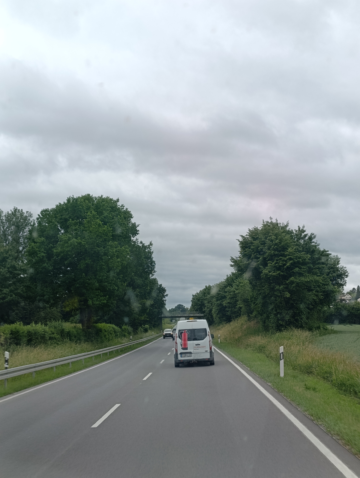 The image depicts a view from a vehicle traveling on a two-lane road, likely a highway, under a cloudy sky. The road surface is asphalt with white lane markings. On the right side of the road, there is a grassy shoulder with wildflowers and a white post with a black stripe. Further to the right, a field of green crops is visible. On the left side of the road, there is a guardrail and a grassy embankment with trees. A white van with "FEUERSCHUTZ Kilmer" written on the back and a fire extinguisher visible through the rear window is in the foreground, in the right lane. A dark-colored car is visible in the distance in the left lane. A bridge or overpass is visible in the background. The environment appears to be rural or suburban, with trees lining the road and buildings visible in the far distance. The overall scene is overcast and potentially humid, as suggested by some smudges on the windshield. The context provided indicates the location is Sigmaringen, Germany.