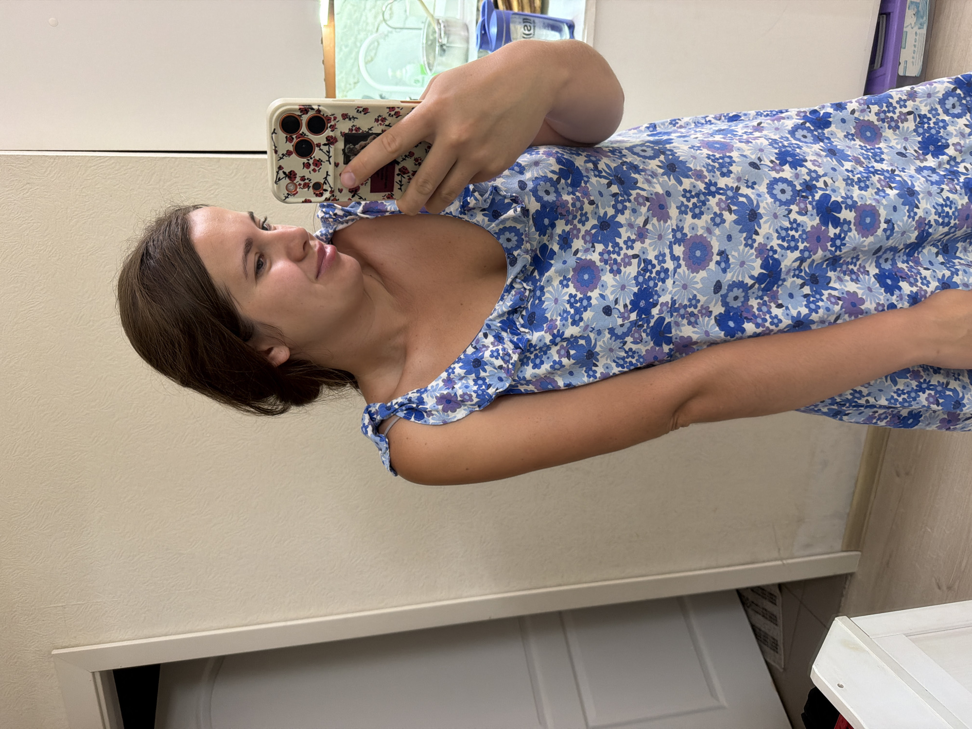 A woman is taking a mirror selfie in a room that appears to be a home. She is wearing a blue floral dress and is holding a phone with a floral case. The room has beige walls, a white door, and wooden flooring. There is a white cabinet in the foreground. In the background, there is a window with a view of what looks like a street or residential area, with glimpses of buildings and possibly some greenery, indicative of an urban setting within a city. Given the context provided, the location is identified as Sochi, Russia.