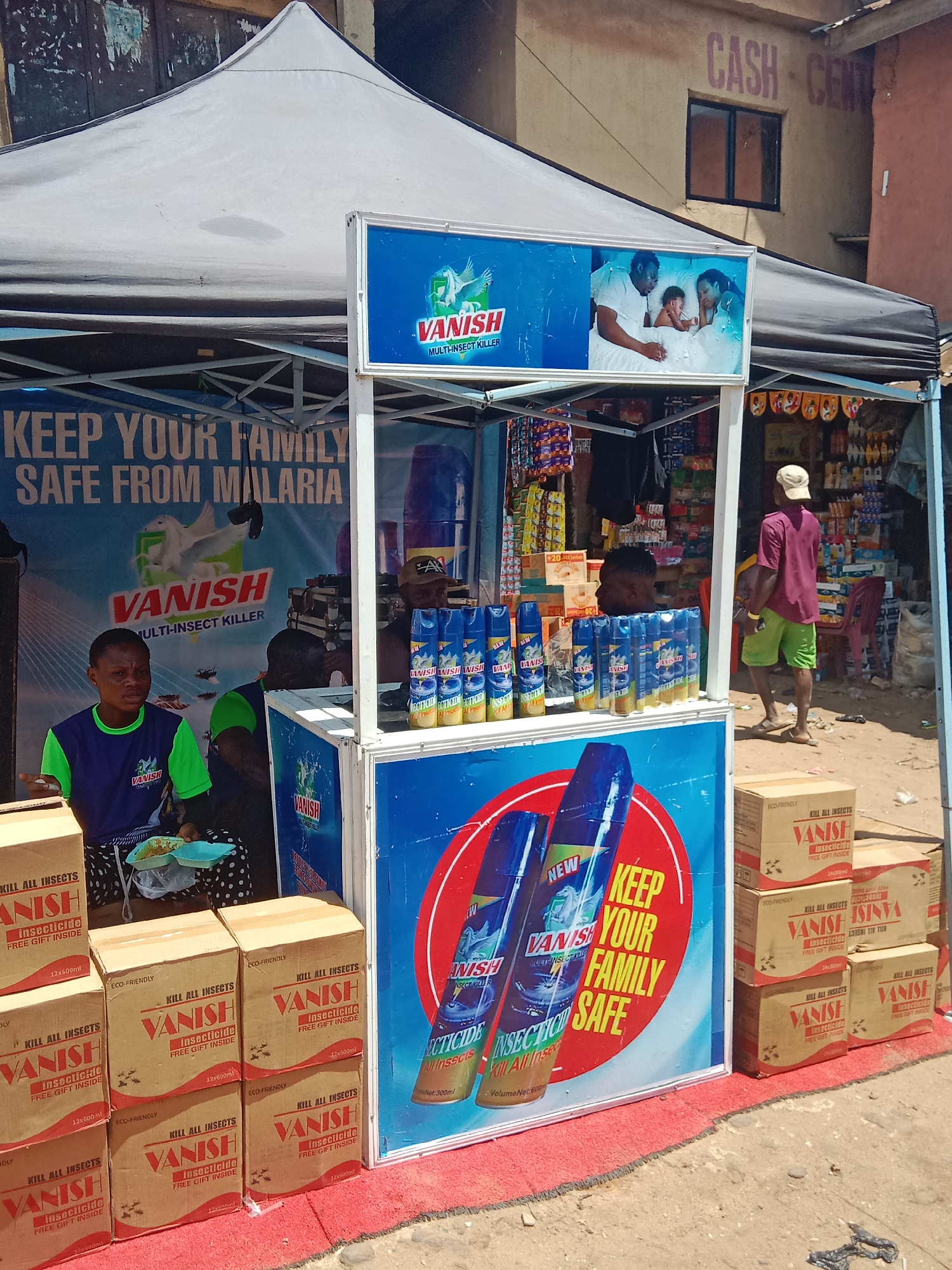 The image depicts a roadside vending stand in Onitsha, Nigeria, selling "Vanish" brand multi-insect killer. The stand, sheltered by a white tent, is staffed by at least two people. A woman is seated on the left, with boxes of Vanish insecticide stacked beside her. A man stands behind the counter, where rows of Vanish aerosol cans are displayed. Signage promotes the product, emphasizing its effectiveness against malaria and its safety for families. The backdrop includes a building with the words "CASH-CENT" visible. A man in a purple shirt and lime green shorts is walking past the stand. The stand itself is decorated with promotional materials. There are various items for sale in the shop next to the stand.